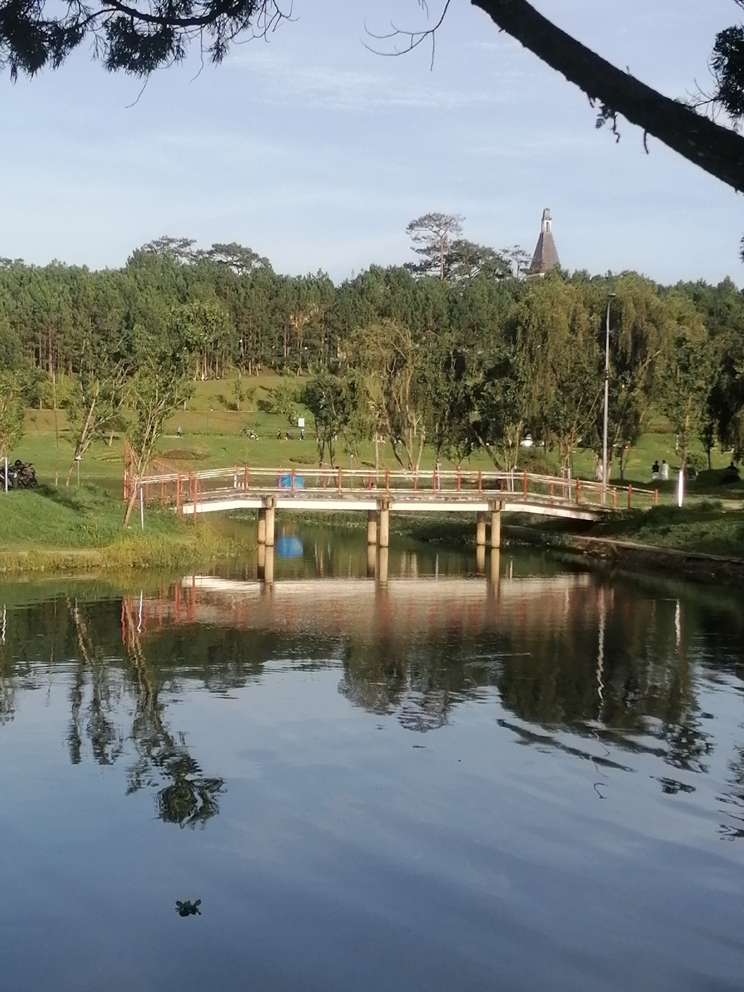 A bridge with white decking and red railings spans across a body of water. The bridge is supported by two concrete pillars. The water reflects the bridge, trees, and a clear blue sky. On the far bank, a grassy slope is dotted with trees and people are visible walking and riding motorbikes. In the background, a forest of pine trees stretches across the landscape, and a distinctive building with a pointed roof and spire is visible above the trees. The scene appears to be a park in the city of Phường Xuân Hương - Đà Lạt, Vietnam.