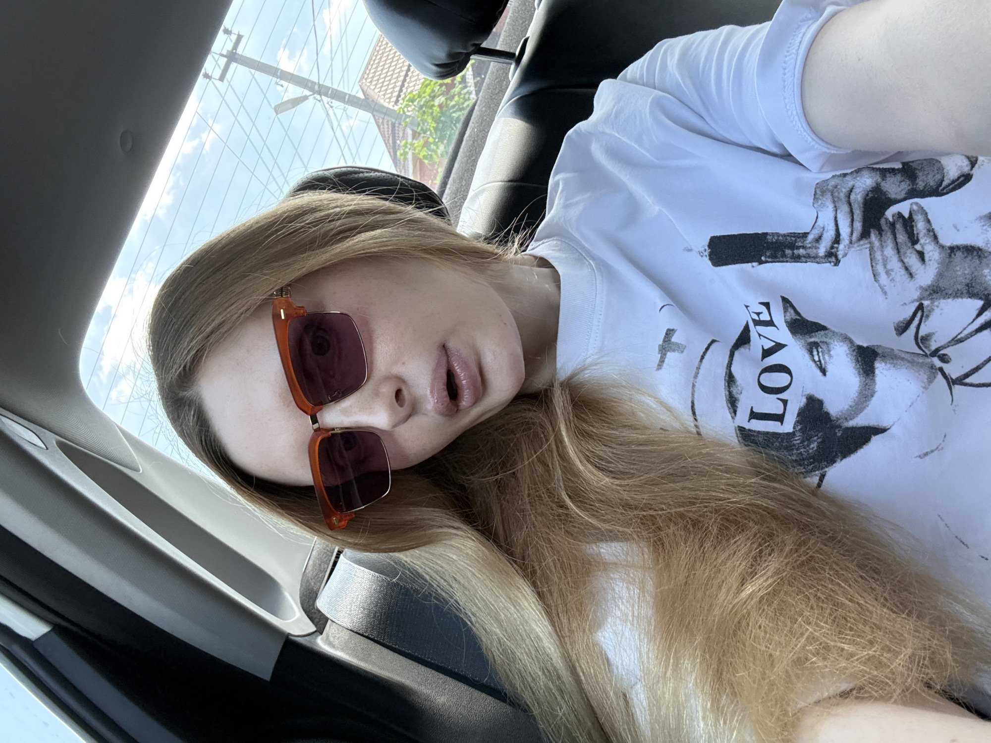 The image shows a young woman with long, blonde hair and sunglasses, leaning back in the passenger seat of a car. She is wearing a white t-shirt with a black graphic design and the word "LOVE" printed on it. Her mouth is slightly open, and her eyes are looking towards the camera. The background shows the interior of the car and the outside through the window, with a bright sky, suggesting it is daytime. The sunlight filtering through the window creates a bright and possibly warm atmosphere. The scene appears to be a casual selfie taken while traveling.