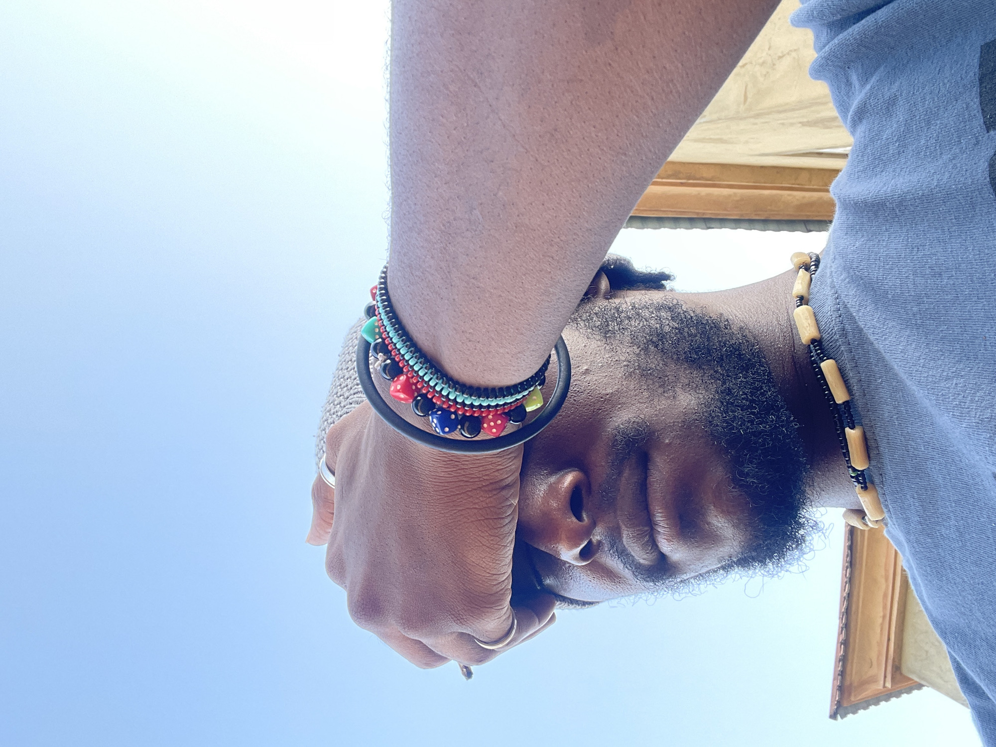 The image is an upward-looking selfie of a Black man with a beard. He is wearing a blue shirt and a beaded necklace. His left wrist has several bracelets, including one with colorful beads and another that resembles dice. He is holding his right hand up near his face, wearing a ring on his right hand. The background is a clear blue sky and part of a building with a wooden exterior. Based on the context, it is presumed that the location is in Jos, Nigeria.