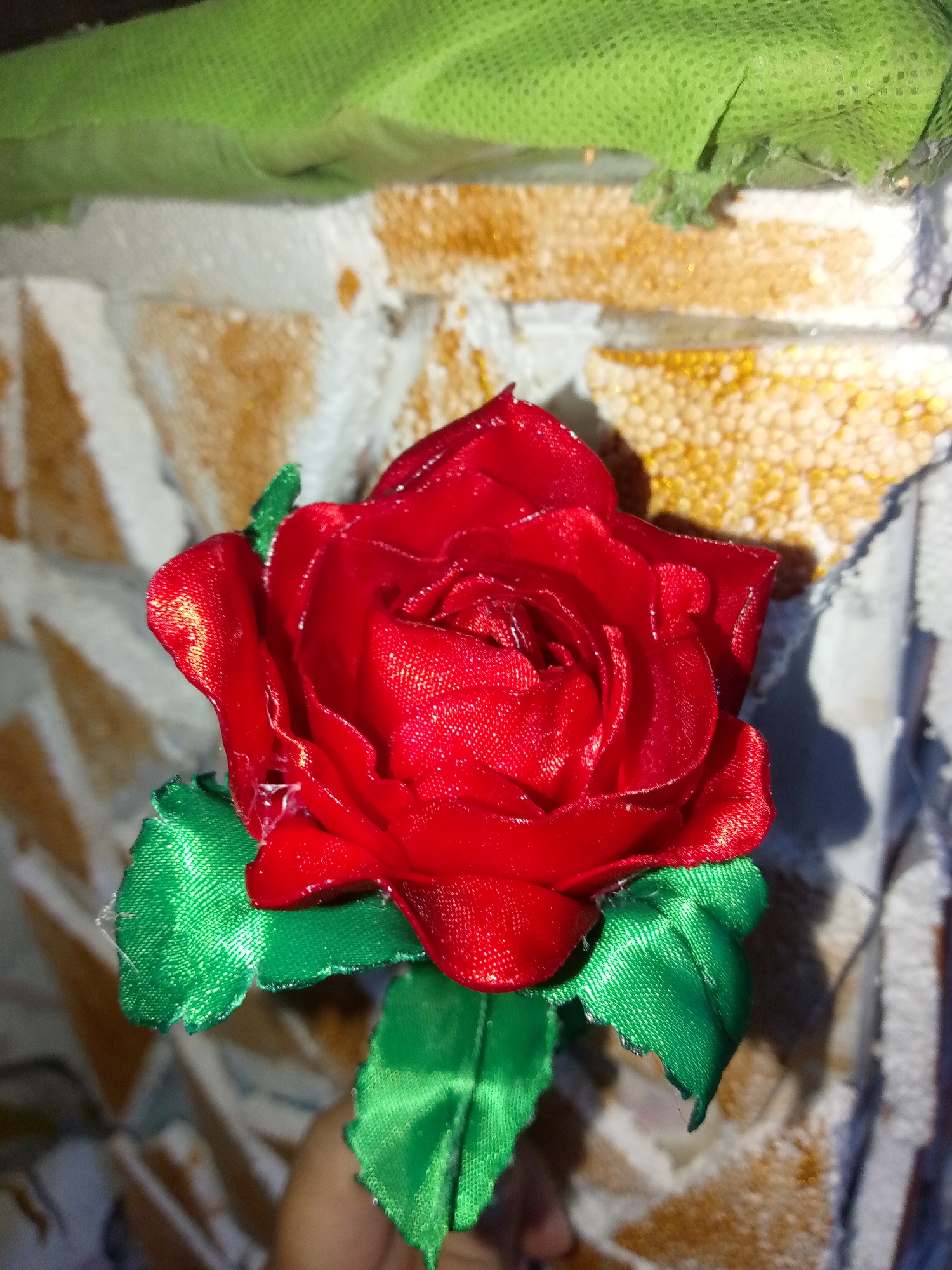 A close-up shot shows a red rose made of fabric with green fabric leaves. The background is a wall with textured, rectangular tiles in light brown and white. In the upper left corner, a piece of green fabric is visible. The lighting suggests an indoor setting. There are no people or other obvious subjects present besides the flower itself. There is no location specific to Juan Griego, Venezuela, discernible from the image.