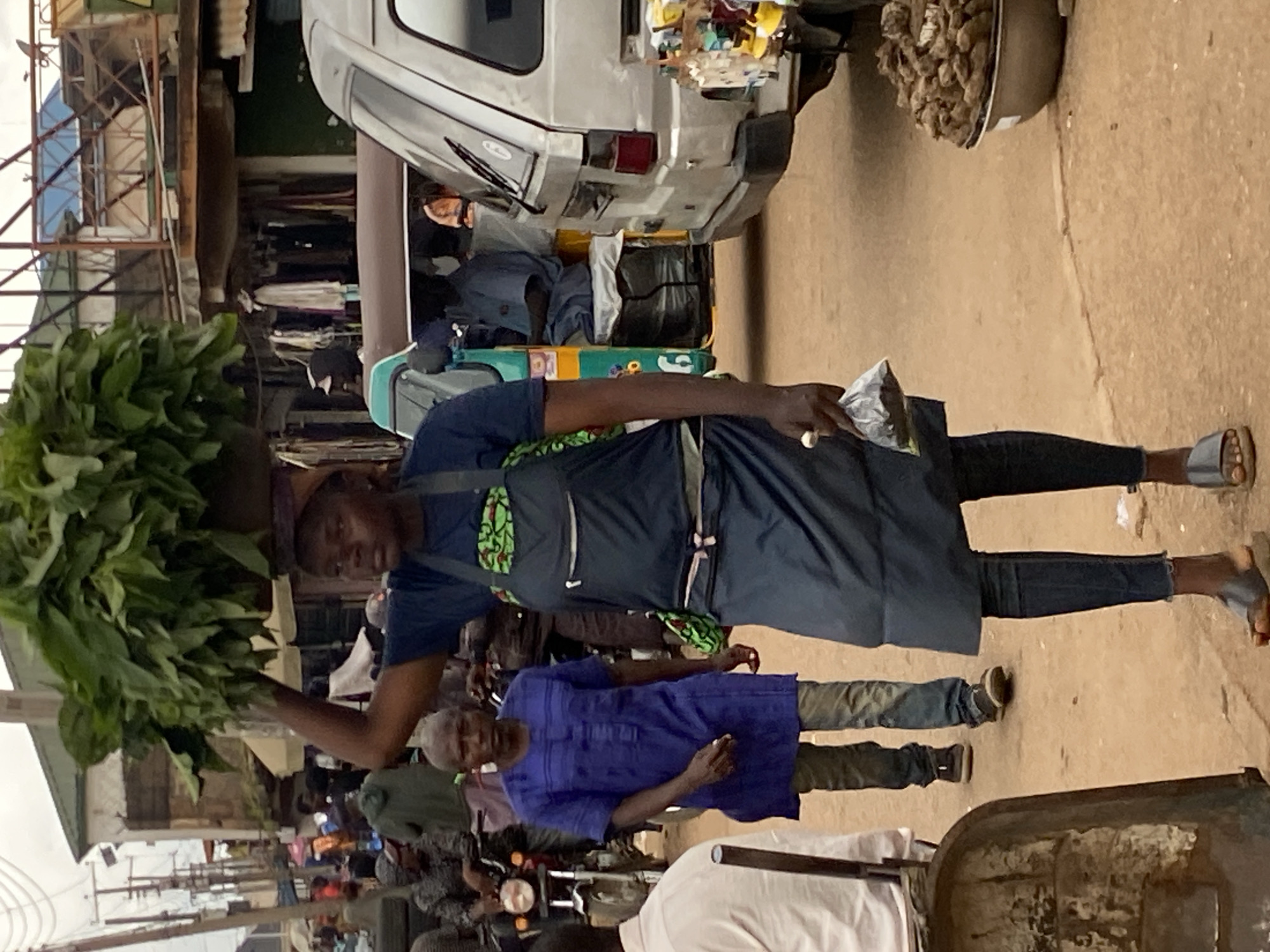 This is a medium shot of a market scene in Bukuru, Nigeria. A woman walks towards the viewer carrying a large bunch of green leafy vegetables on her head, balanced with a cloth. She is wearing a dark blue t-shirt with a green and white patterned apron over it, and dark blue jeans. Her left hand holds a small plastic bag filled with a dark substance. Beside her, to her left, a man in a bright blue tunic and dark pants walks away from the viewer. Behind them, various market stalls and vehicles are visible, including a silver van, a green and yellow auto-rickshaw, and several motorcycles. The ground is unpaved and dusty. In the foreground, to the left, the edge of a weathered metal cart is visible. To the right, a large metal bowl filled with brown, root-like objects sits on the ground.