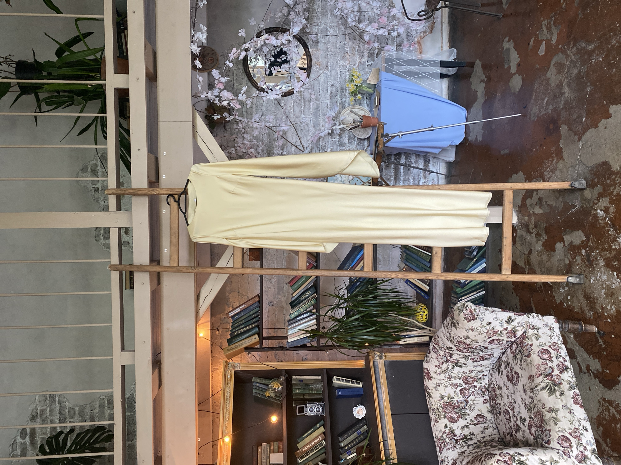 The image depicts an interior, likely a shop or studio, in Yekaterinburg, Russia. A cream-colored dress hangs on a wooden ladder. Behind the ladder, there is a wall with bookshelves containing numerous books, some plants, and an ornate mirror. A floral armchair is positioned near the base of the ladder on the right. The floor is a mottled mix of brown and gray. A studio lighting rig with a blue backdrop sits in the background. The overall scene gives a Bohemian, vintage aesthetic.