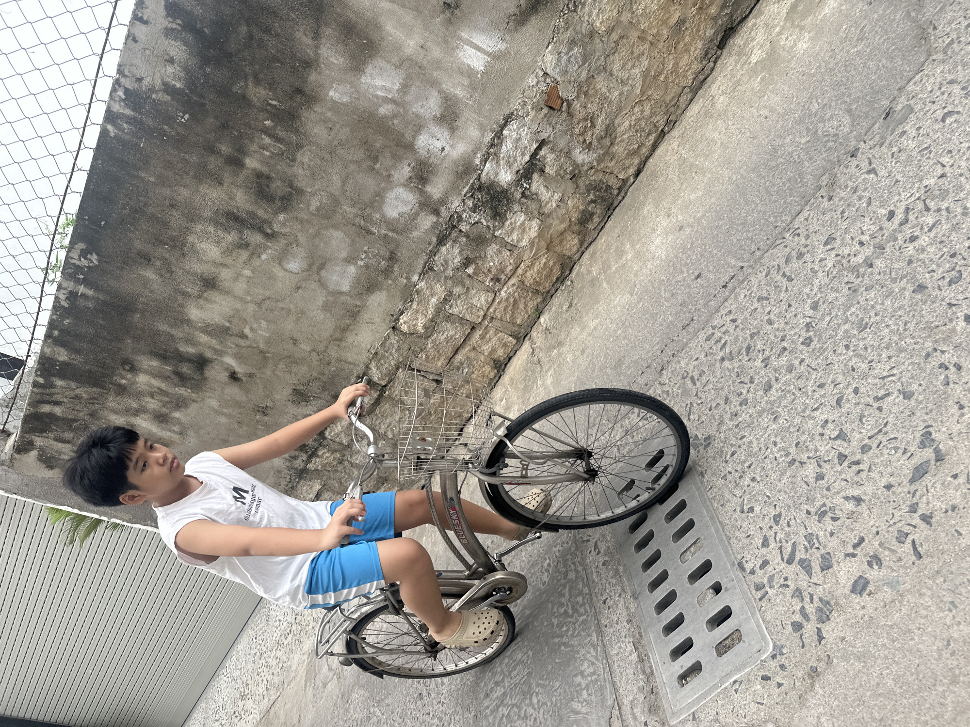 A boy is sitting on a bicycle in Bac Nha Trang Ward, Vietnam. The background shows a concrete wall topped with a chain-link fence, as well as a concrete sidewalk. The bicycle is silver with a basket on the front. The boy is wearing a white tank top and blue shorts, and he is wearing beige sandals on his feet. The front wheel of the bicycle is next to a concrete grate. The image is taken at an angle.