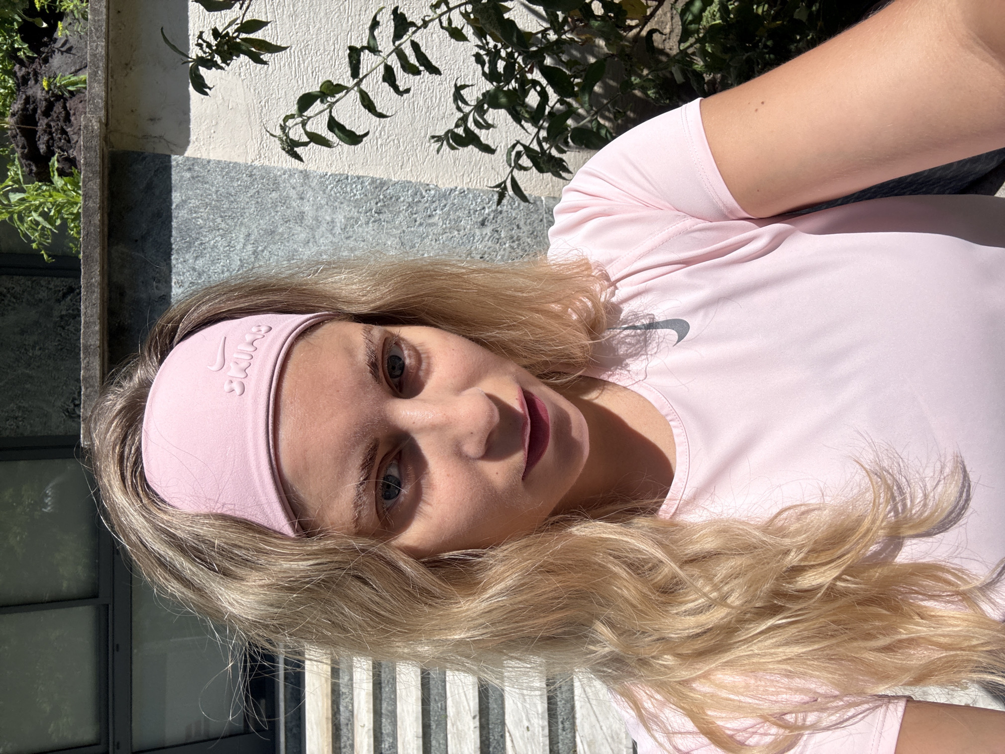 A young woman is pictured wearing a pink Nike headband and pink shirt. She is outside and in the sunlight, next to a white wall with a plant and some greenery on top of the wall. There is what looks like a wooden bench behind the woman. It is not possible to tell if she is in the city of Paris.