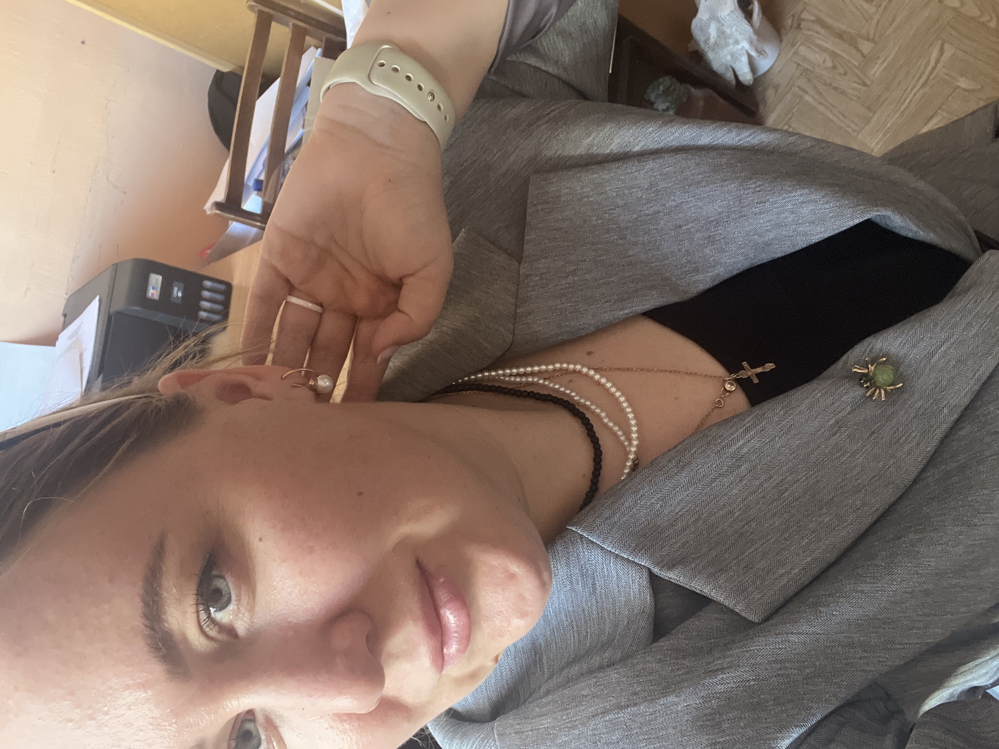 A woman is taking a selfie indoors. She wears a grey blazer over a black top and several necklaces: one made of black beads, a pearl necklace, and a gold chain with a cross pendant. She also wears a white smart watch and hoop earrings with pearls. A green brooch is pinned on her blazer. A printer is visible in the background, and a portion of a wooden chair is also visible. The location is likely indoors, but the specific city of Chaikovsky, Russia, cannot be determined from this image.