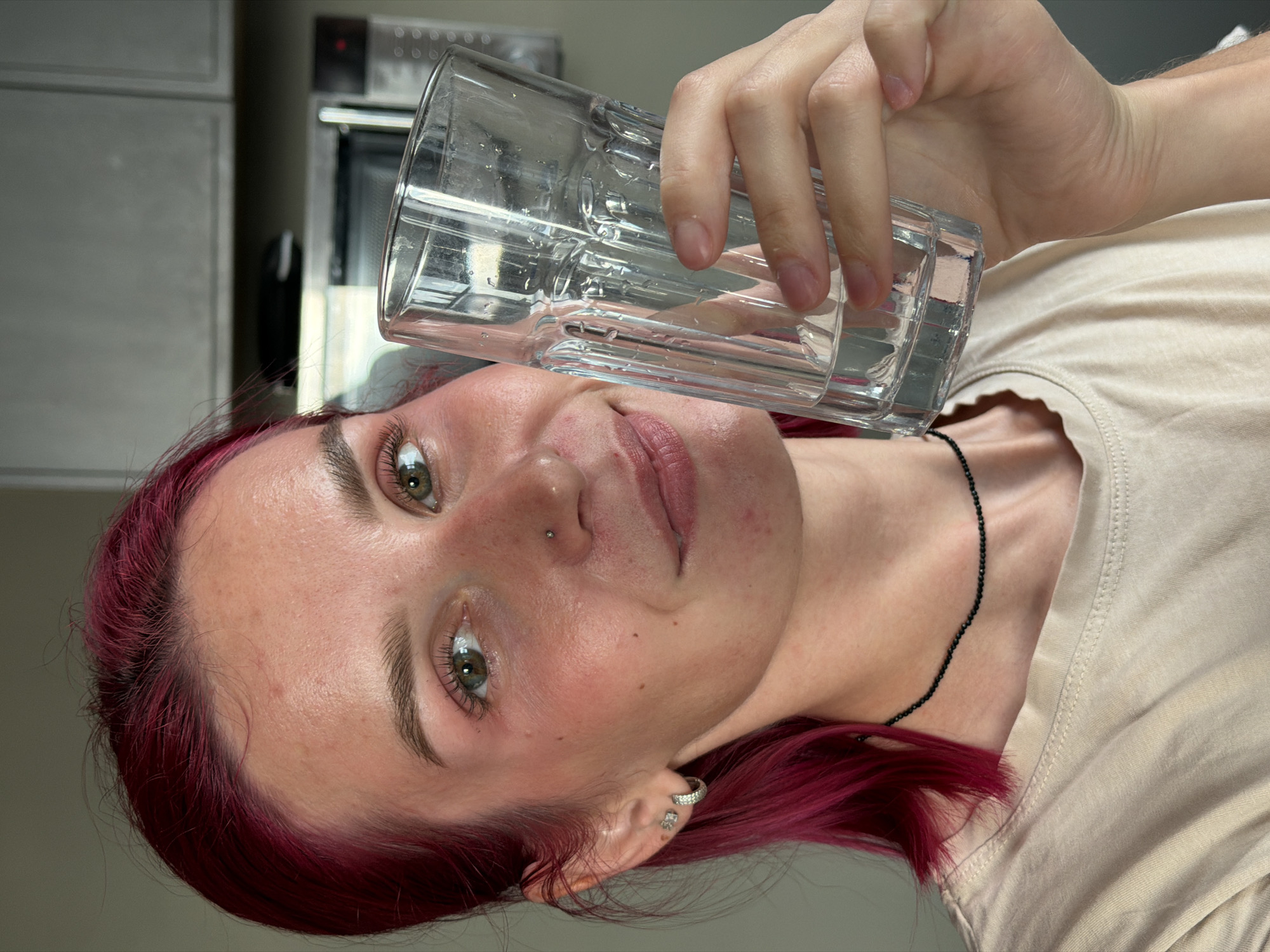 The image shows a close-up, upside-down view of a young person with vibrant magenta hair holding a glass of clear liquid. The person has light-colored skin with some redness on the cheeks, green eyes, and prominent eyebrows. They are wearing a light-colored t-shirt and a thin black beaded necklace. A silver earring with a small clover-shaped charm is visible in their left ear. There is also a small metal stud piercing on their nose. The background appears to be a kitchen, with what looks like a microwave or oven visible on the left side. The lighting suggests it is daytime, and there are no indicators of weather conditions. The overall mood is casual and candid, with the person looking directly at the camera. No visible text is present in the image. The location context provided is Chaikovsky, Russia.
