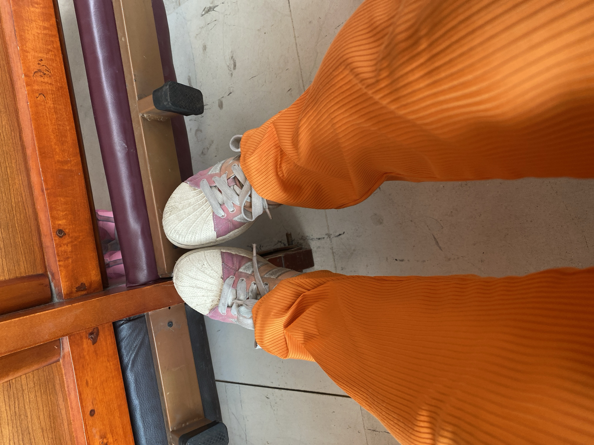 A top-down shot shows a person wearing orange, horizontally ribbed pants and white and pink sneakers, standing on a light-colored tile floor. The left side of the frame includes part of a wooden bench or structure with a dark brown frame, a burgundy cushion, and a black foot. No people are fully visible, only the lower portion of a person wearing orange pants and sneakers. The setting appears to be indoors with basic tile flooring. The photograph shows a view of the floor, the lower part of someone in orange pants and pink shoes, and part of a wooden bench/structure on the left. There is no visible information indicating that the photo was taken in the city of San Bernardino Tlaxcalancingo, Mexico.