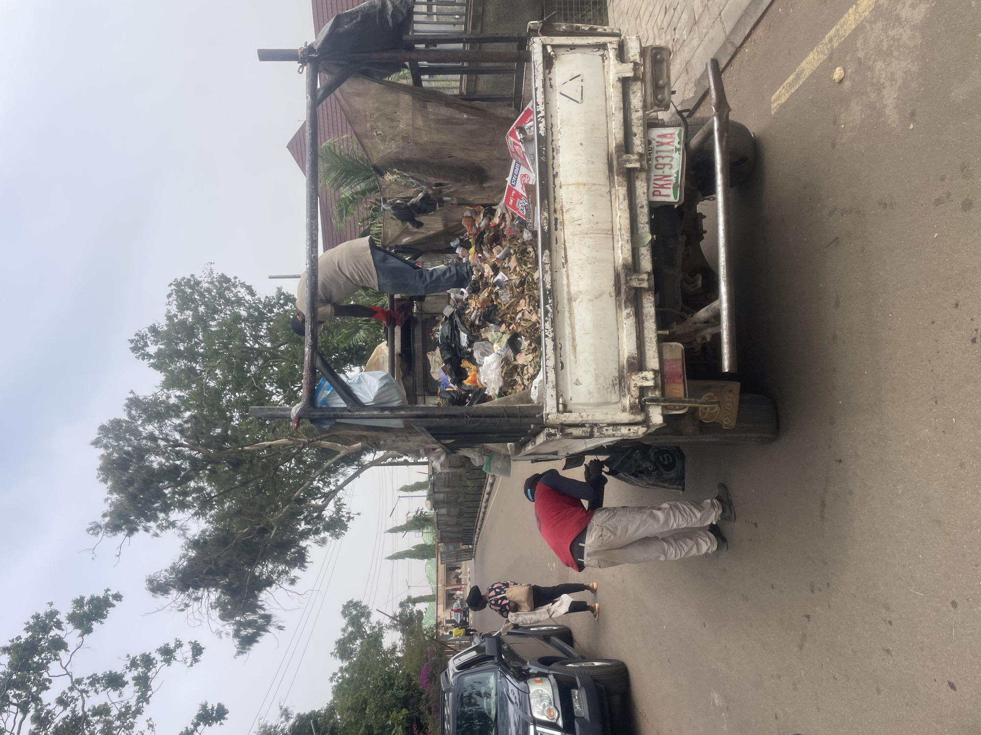 The image shows a sanitation crew loading garbage into the back of a white truck on a paved street in Jos, Nigeria. The truck's license plate reads "PKN-931XA". One sanitation worker is inside the truck bed, tossing garbage further in. Another worker is on the ground, picking up trash bags. A black SUV is parked behind the sanitation truck. A tree overhangs the street. Other people are visible in the background along the sidewalk.