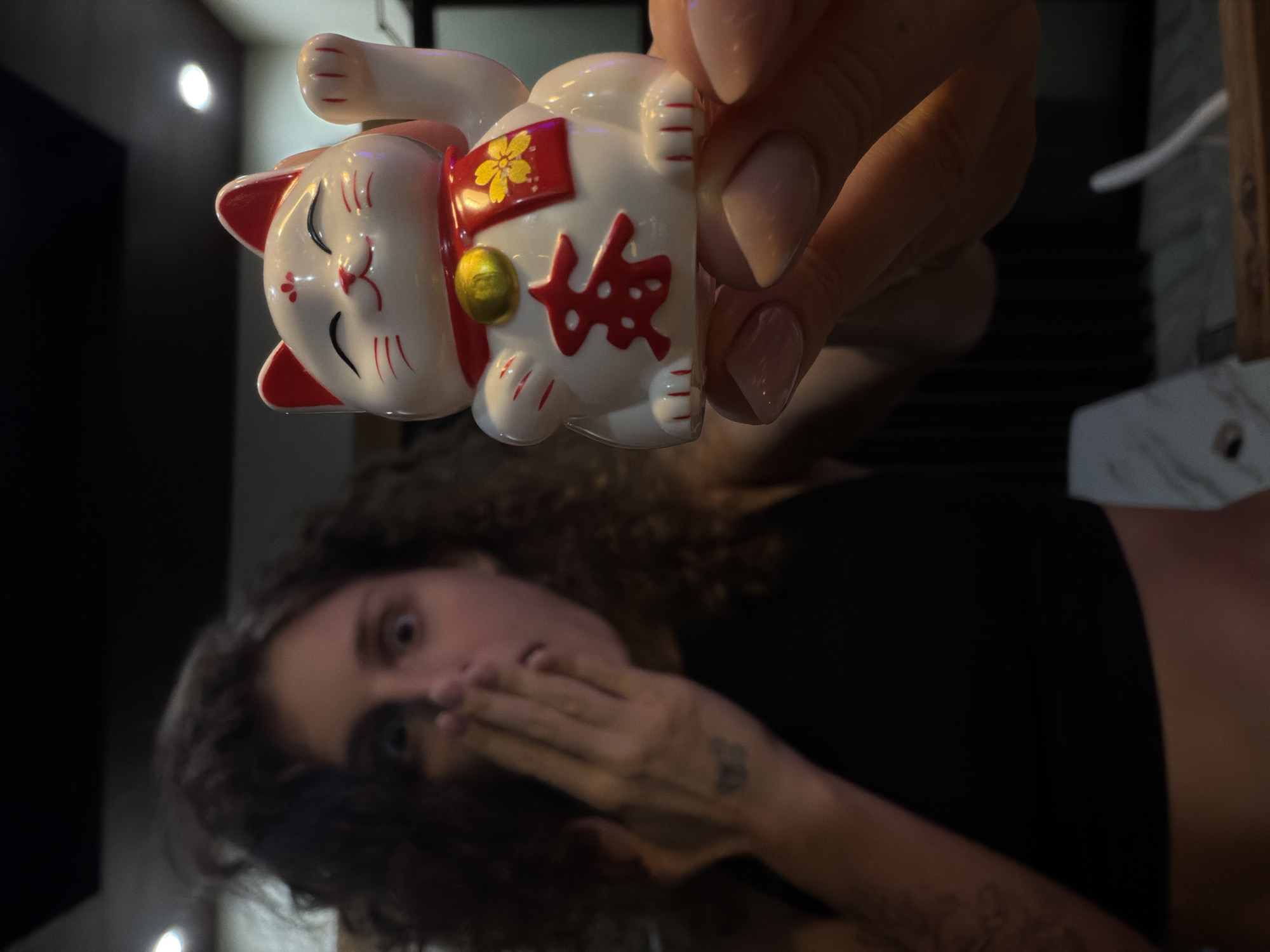 An indoor, low-light shot depicts a hand holding a white Maneki-neko (lucky cat) figurine in the foreground, with a person reacting with surprise in the blurred background. The scene is set in North Kuta, Indonesia.
The ceramic Maneki-neko is predominantly white with red details, including its ears, whiskers, and paw outlines. It wears a red collar with a golden bell and a red bib adorned with a yellow floral pattern. On its body, a prominent red Japanese kanji character, likely "福" (fuku, meaning good fortune or luck), is visible. The cat's right paw is raised, a traditional gesture for inviting money or good fortune. The hand holding the figurine has neatly manicured, light-colored nails.
In the blurred background, a person with curly dark hair and wide-open eyes expresses surprise or playful shock, covering their mouth with one hand. They appear to be wearing a dark, possibly black, top. A faint tattoo can be seen on their visible forearm. The overall lighting is dim, with a few bright, circular light sources in the distant background suggesting an indoor, possibly evening, setting. The primary focus is on the Maneki-neko and the hand holding it.