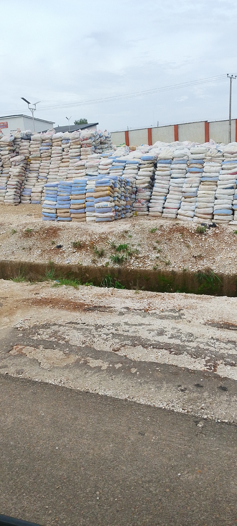 This media file depicts a large outdoor storage area filled with numerous sacks of cement. The sacks are stacked high, forming towering piles. Some sacks are white, while others are blue and yellow, indicating different types or brands of cement. In the background, a low-rise building with a white facade and a dark roof is visible, along with a fence topped with barbed wire. Solar-powered lights are mounted on poles near the building. The ground in the foreground is a rough, uneven surface of asphalt and gravel, with a concrete ditch running through it. The sky is overcast, suggesting a cloudy day. No people are visible in the image. The overall scene appears to be a storage yard for construction materials. Some visible text on the sacks includes "BUA P.O.P. 42.5", "LAFARGE CEMENT", and other unreadable markings.