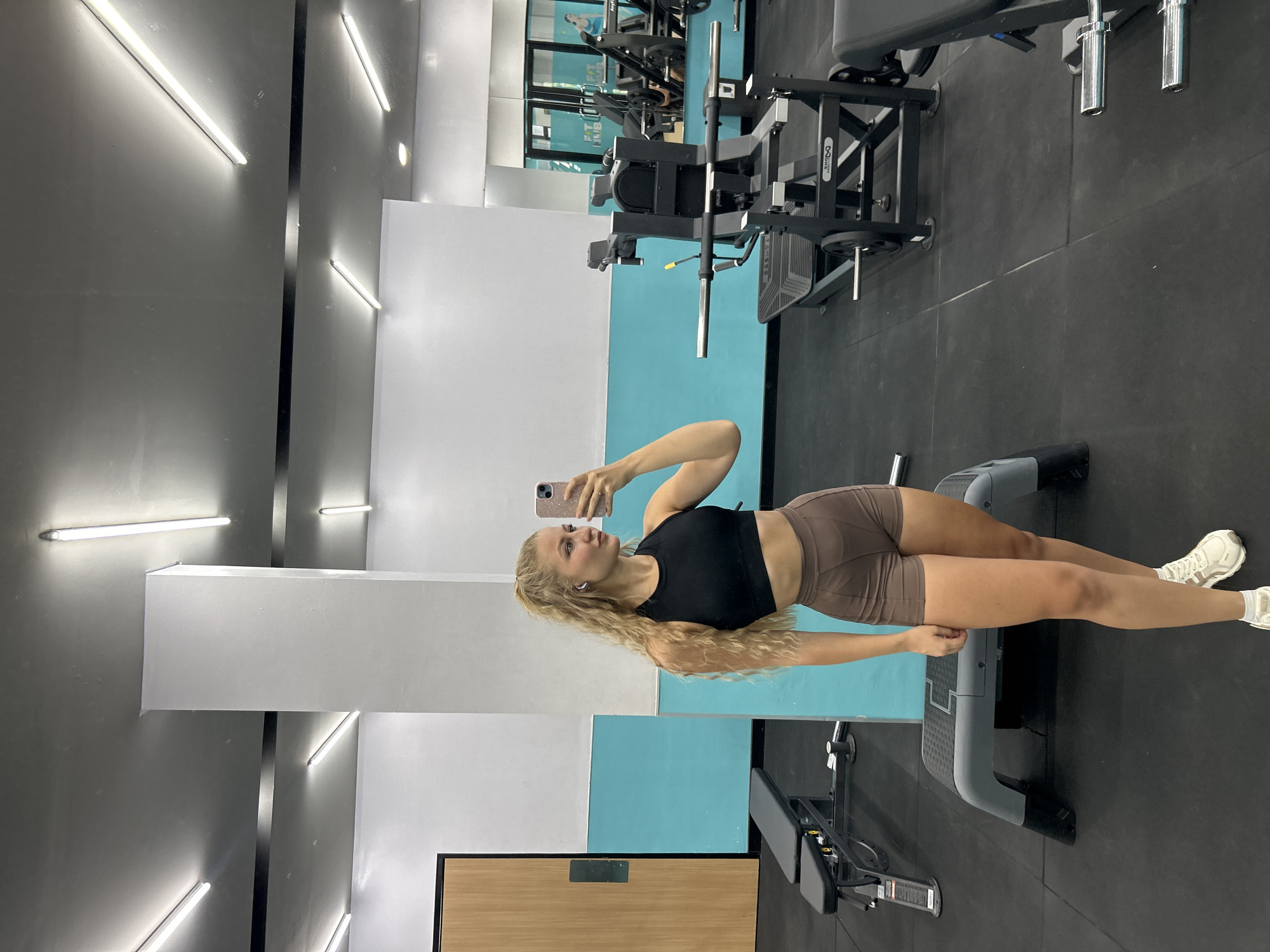 A young woman with long, wavy blonde hair is captured taking a selfie in a gym setting. She is wearing a black sports bra, brown athletic shorts, and white sneakers, holding a pink smartphone. She appears to be wearing wireless earbuds. Her expression is focused, looking into her phone as she holds it up.
The scene is an indoor gym, characterized by a contemporary design with walls painted in sections of black, white, and a vibrant light blue. Bright, horizontal LED strip lights illuminate the space. Various pieces of dark-colored gym equipment are visible, including a weight bench with the legible brand "OSSO" on its side, and what appears to be a large leg press or multi-station machine in the background. Mirrors on the walls reflect parts of the gym, showing more equipment and a faint, mirrored text on a blue wall, possibly reading "FIT KIDS" or similar. The floor is covered with dark rubber matting. A light wooden door with a black frame is partially visible at the bottom left of the frame. The overall impression is a modern, well-lit fitness facility in Kuta Selatan, Indonesia.