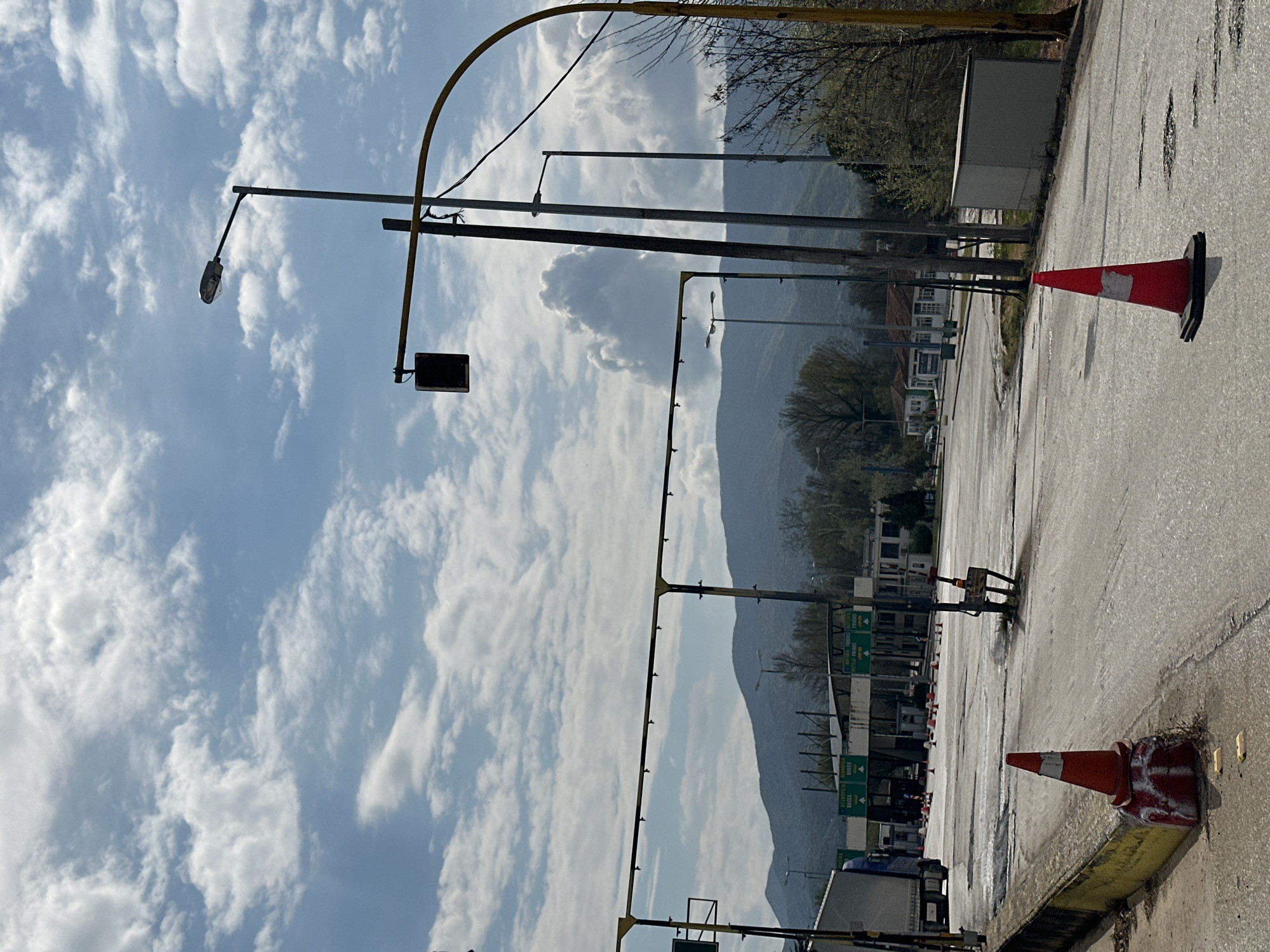 The image shows a highway toll booth in the Δημοτική Ενότητα Προμαχώνος, Greece. The scene includes multiple toll lanes with signage in Greek. Two traffic cones are present in the foreground: one on the far right and one near the bottom of the frame. Several light poles are positioned along the highway, as well as an arch-shaped structure over the lanes. The sky is partly cloudy. A mountain range appears in the background. The asphalt road surface is visible.