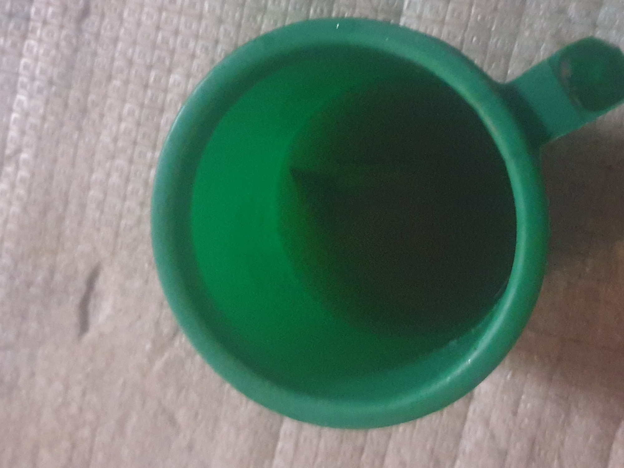 A close-up, overhead shot captures a green plastic cup filled with a dark liquid, possibly coffee or tea, resting on a textured, beige surface. The cup has a handle extending from its right side. The surface appears to be a placemat or tablecloth with a subtle grid pattern. The lighting is dim, and the focus is on the cup, with the background slightly blurred.