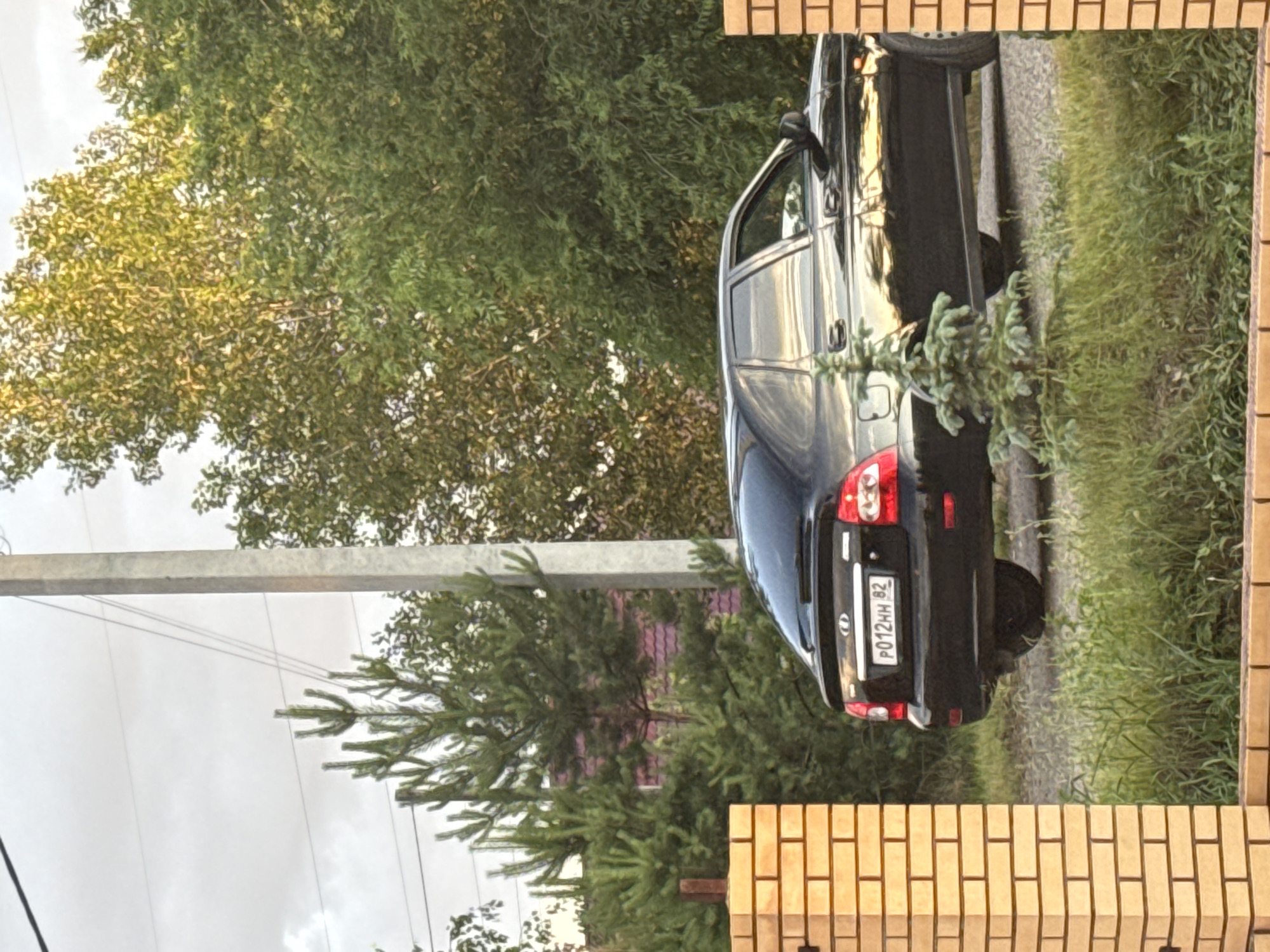A black car is parked on a gravel road next to a fence made of light brown bricks. The car is a sedan and has a license plate that reads "P012HH 82". Trees with green leaves surround the scene. The sky is overcast. The image appears to be taken in the afternoon or evening. The location is Minusinsk, Russia.