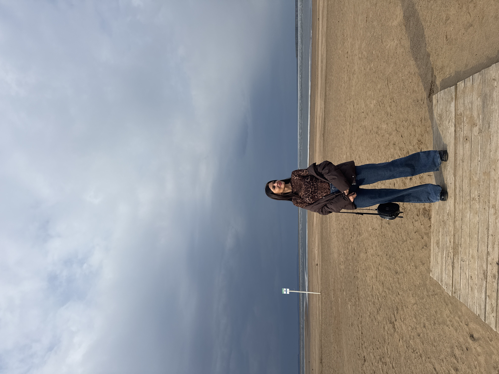 A woman stands on a beach near Chaikovsky, Russia. She is wearing jeans, a dark jacket, and a leopard-print shirt. A purse rests on the wooden planks beside her. The sky is overcast, and the beach stretches out to the horizon. A small sign is visible further down the beach. The scene appears to be a candid snapshot of a casual day at the beach.