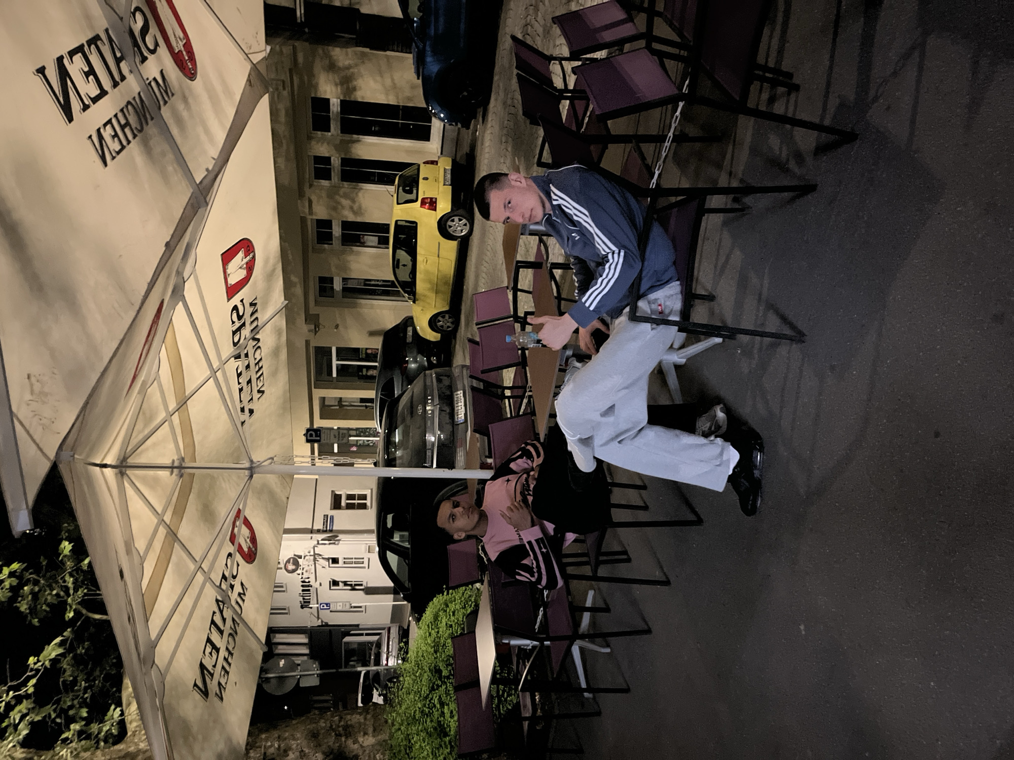 The image shows two individuals sitting on chairs outside, likely at a café or bar, under an umbrella. The first person is a young man wearing a blue and white tracksuit and light-colored pants, holding what appears to be a plastic water bottle. The second person is wearing a pink and black shirt with black pants. Several dark-colored chairs are arranged around them. Behind them is a street scene with parked cars, including a yellow and black one, and buildings with lit windows. The location is in an urban area.