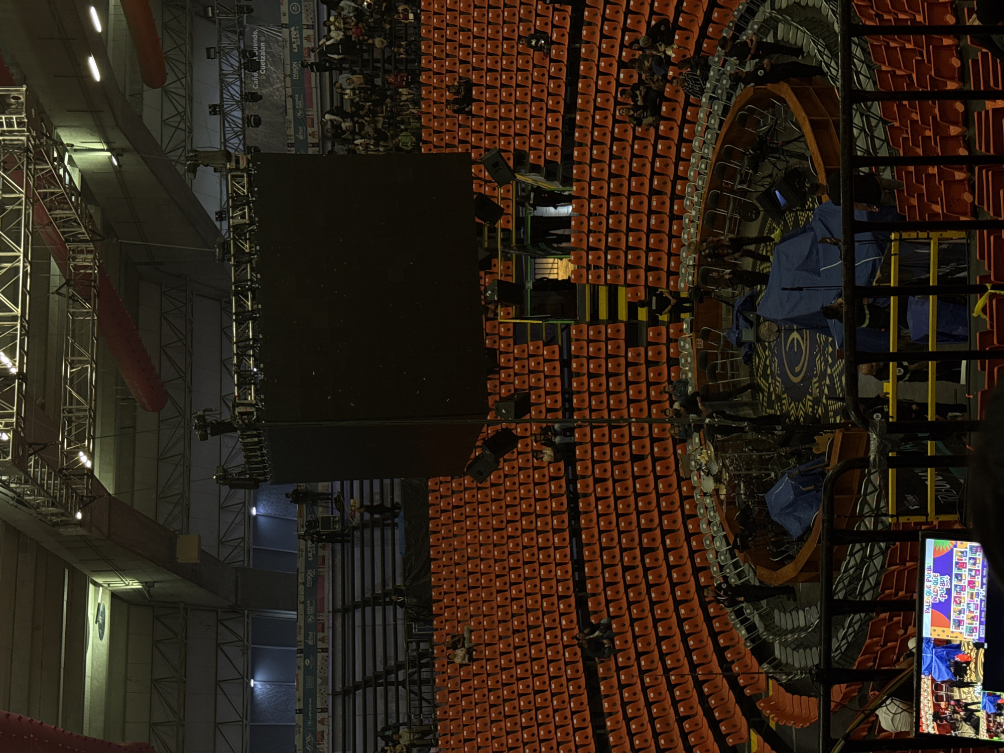 The image captures a view of the Palenque de la Feria de Puebla in Puebla, Mexico. The arena is filled with rows of orange seats, mostly occupied by people. A large black screen dominates the central area. There is lighting and rigging above the stage. A tiered seating arrangement surrounds the stage. A screen in the foreground displays event information and imagery of the crowd.