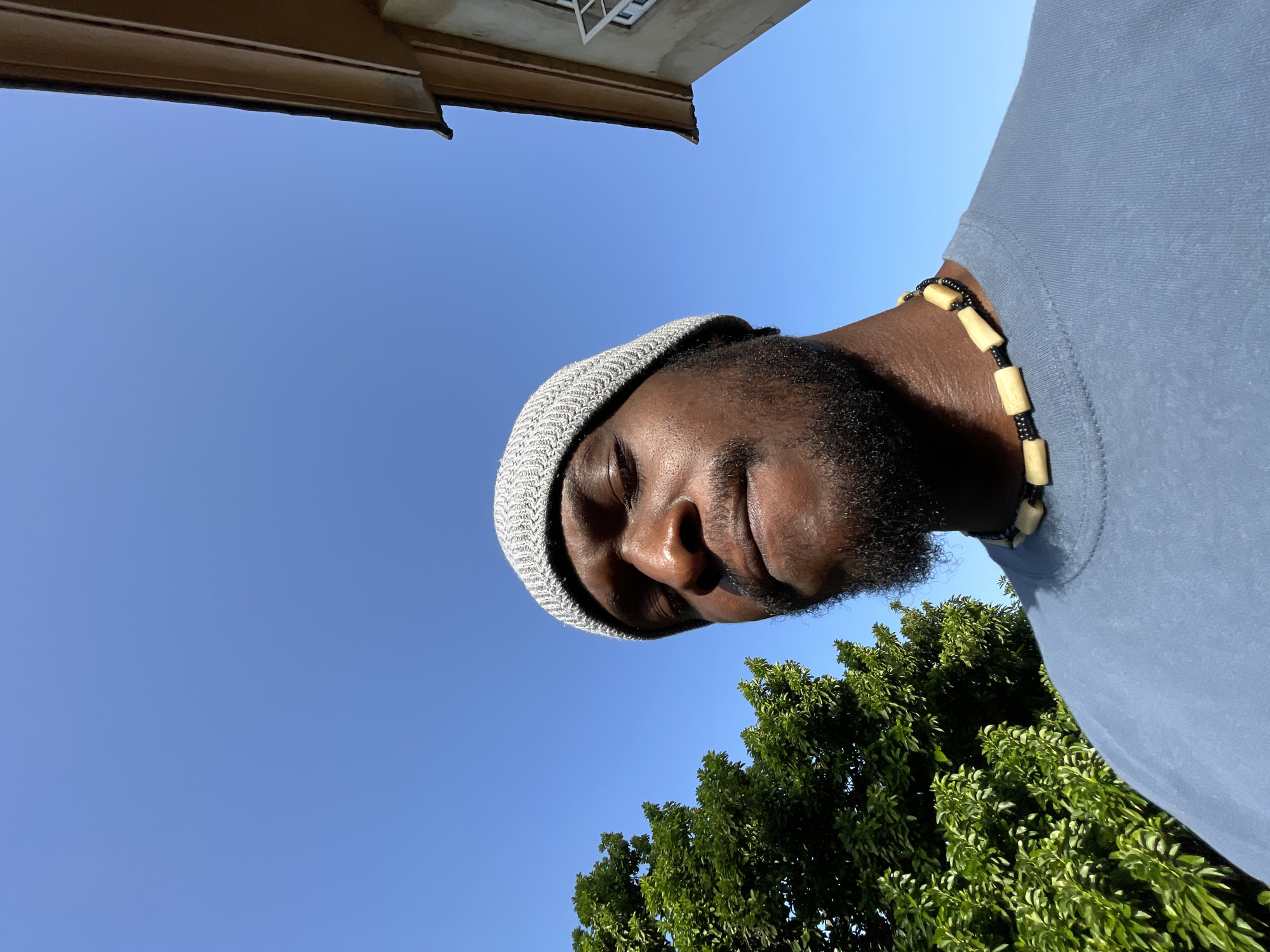 The image is an upward-angled selfie of a Black male wearing a gray knit hat and a light blue t-shirt. He also has a beaded necklace. He is outdoors under a clear blue sky. The top of a building is visible at the upper left, and green trees are at the lower right. The location is not discernable as being in the city of Jos, Nigeria from the image alone.