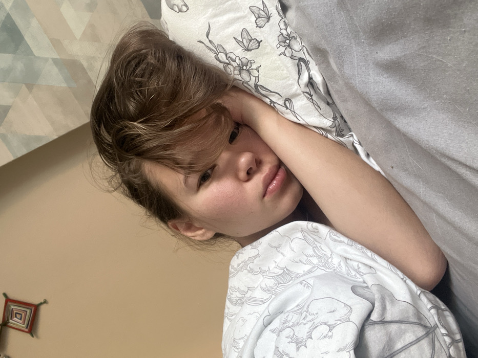 A young woman is resting in bed, propped on pillows. Her blonde-brown hair is messy, with bangs falling across her forehead. She gazes directly at the camera with a slightly pensive expression. She is wearing a white shirt with a whimsical print. The bedding includes patterned pillows and a grey blanket. A wall with geometric art is visible in the background, along with a small, colorful tapestry. The image is shot at a slightly tilted angle.