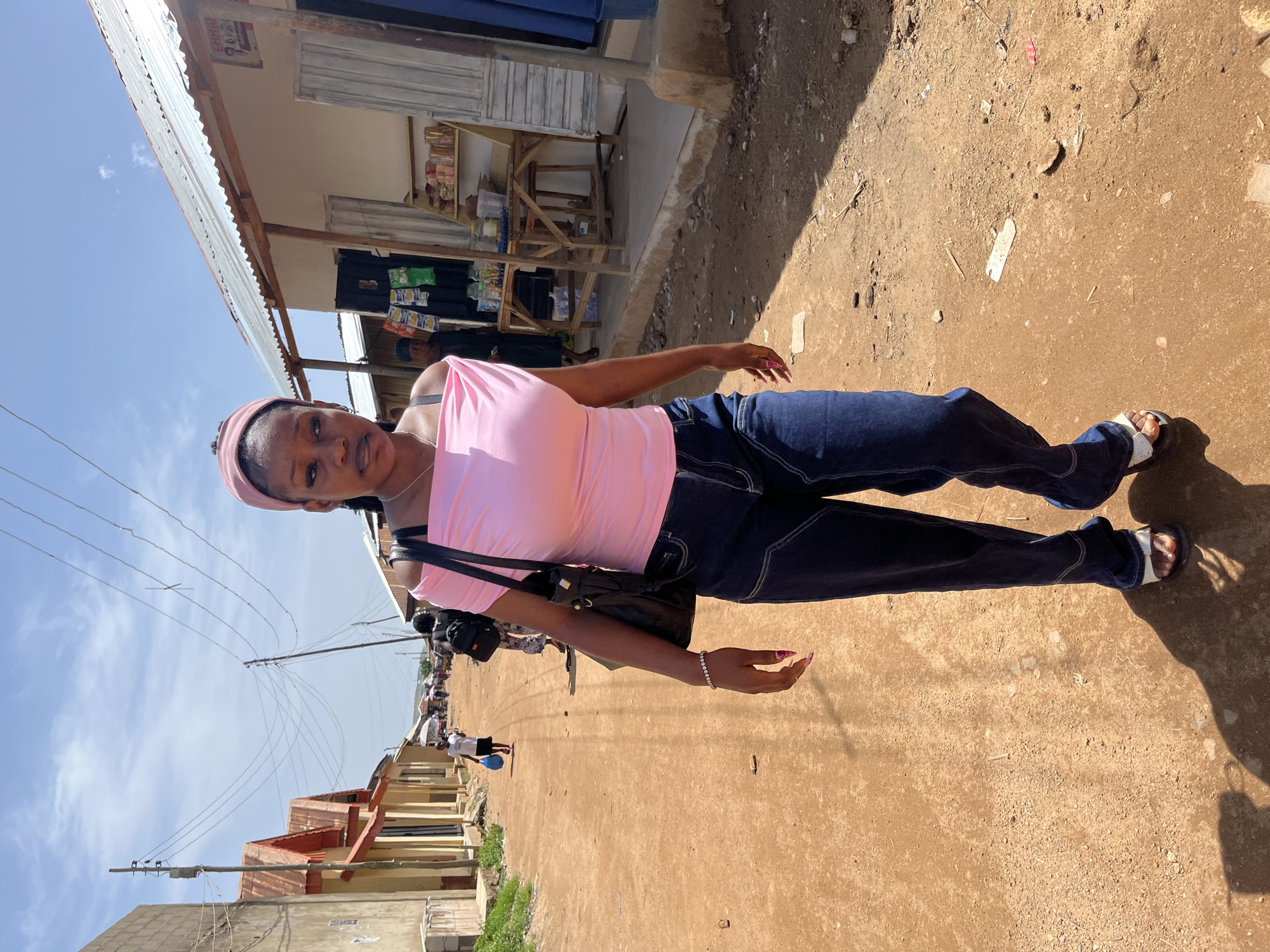 The image captures a young woman standing on a dusty, unpaved street in Jos, Nigeria, under a bright, sunny sky. The central figure faces the camera, wearing a light pink sleeveless top, dark blue jeans with white stitching, and white sandals. A pink headband or headscarf complements her outfit, and she carries a black shoulder bag. A delicate bracelet adorns her wrist, and her fingernails are painted.
Behind her, to the right, is a simple building with a corrugated metal roof, likely a shop, displaying various items on wooden shelves. Above the shop's entrance, a sign is visible, but the text is largely indistinct, possibly starting with "EMB". The street, made of reddish-brown earth, extends into the background, where other people can be seen walking or congregating. Numerous power lines crisscross the blue sky, connected to wooden utility poles that line the street. Strong shadows cast on the ground indicate a mid-day or afternoon setting. The overall scene portrays a typical bustling street environment in a local community.