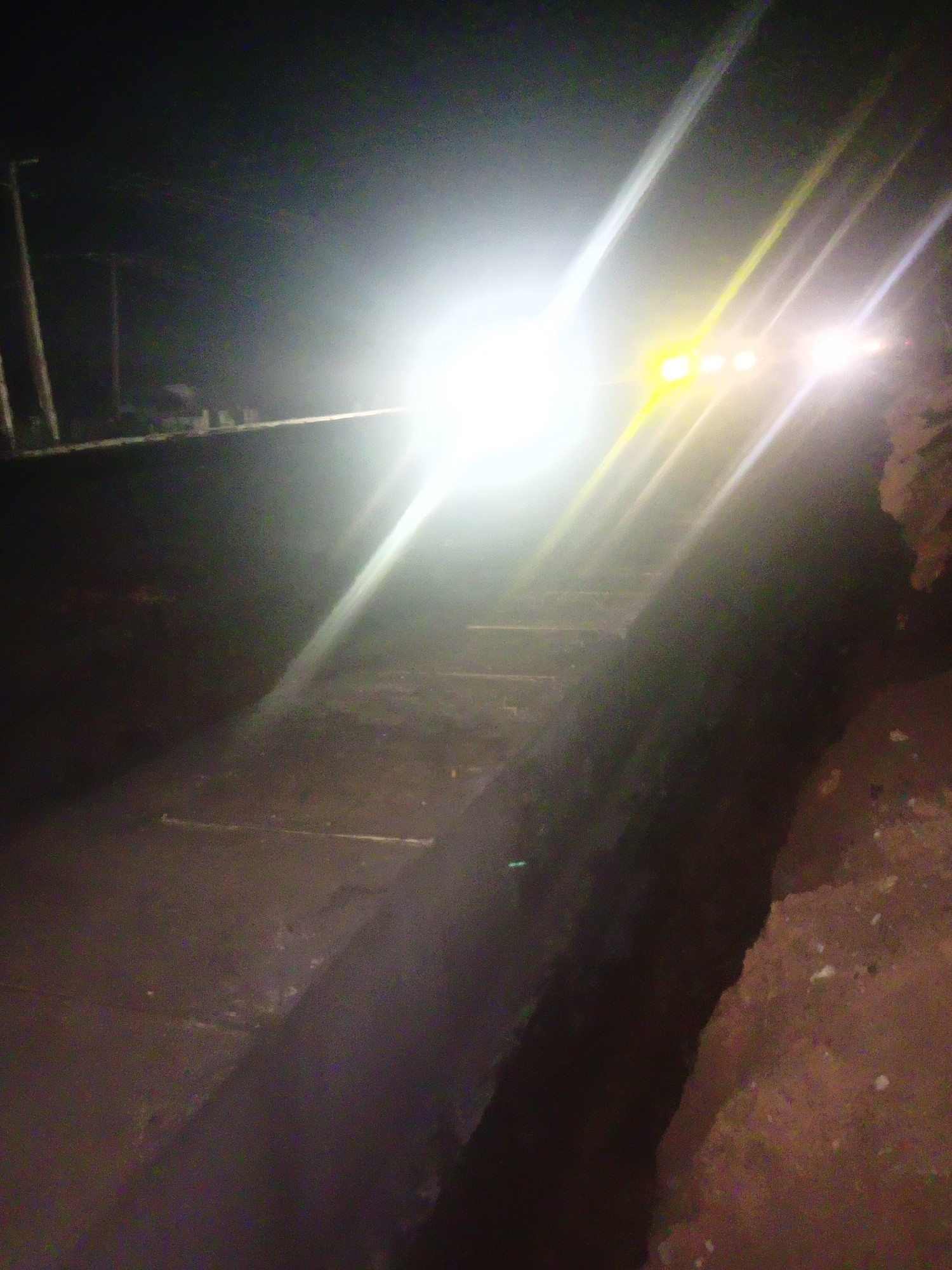The image depicts a dark, outdoor scene, likely at night, with a prominent bright light source emanating from the upper center. This light, possibly from a vehicle's headlights, creates strong glare and lens flare effects, obscuring details in the background. In the foreground, a concrete structure, possibly a drainage channel or a low wall, runs diagonally across the frame. The texture of the concrete appears rough and worn. The ground is dark and uneven, with some scattered debris or rough surfaces visible on the right side. In the far background, faint vertical structures resembling utility poles can be seen against the dark sky, suggesting a semi-urban or rural environment. The overall impression is one of low visibility, with the intense light dominating the scene and making it difficult to discern specific details of the surroundings or any potential occupants of the area. The location in Kakuri Gwari, Nigeria, implies a possibly dusty or unpaved environment, consistent with the rough textures and darkness observed.