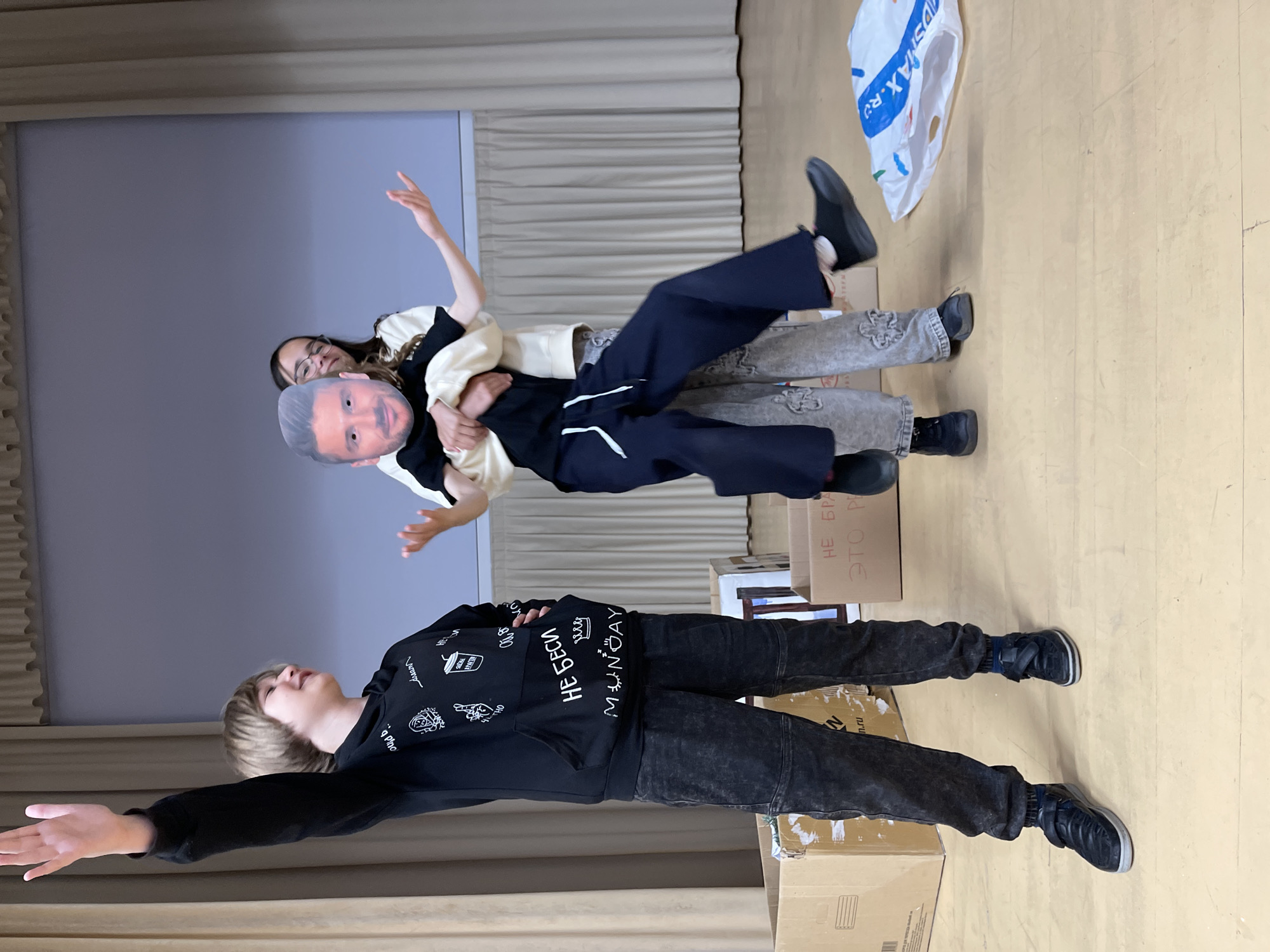 In a brightly lit room, possibly a school or community center, three individuals are engaged in a playful interaction. A young boy with light brown hair stands to the left, raising his right hand in a wave or gesture of greeting. He wears a black hooded sweatshirt with white graphics and dark, textured pants, and black sneakers. In the center, two other individuals are intertwined. One, appearing to be a young woman, is being held from behind by another person. A cutout image of a man's face is held up, obscuring the face of the person in the middle. The person in the middle wears a light-colored shirt over a black t-shirt and patterned gray pants. The person behind them wears dark blue pants. The room has a light-colored wooden floor and beige curtains in the background. Cardboard boxes are present on the floor, one to the left and another in the middle with Russian text that appears to read "НЕ БРА" and "ЭТО Р". A white plastic bag with colorful markings is to the right. The overall atmosphere suggests a lighthearted, perhaps theatrical, moment. The location is identified as Zelenogradsk, Russia.