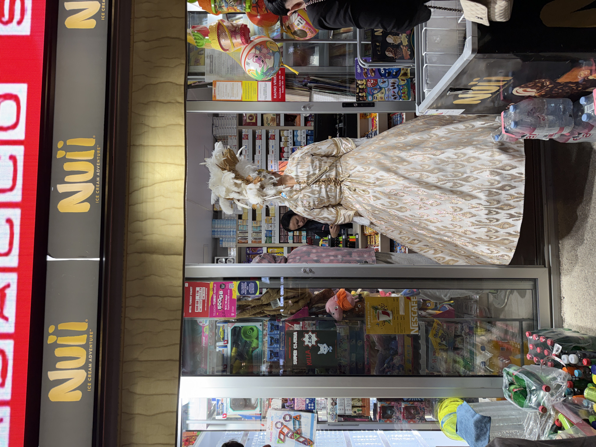 Here is a forensic description of the image:
The image shows a person in a white and gold patterned formal gown and a large feathered headdress standing inside a small retail store or kiosk. The store appears to be in Budva, Montenegro, based on the visible signage in the local language. The person is positioned between shelves stocked with various products, including snacks, drinks, toys, and promotional telecom materials. Signage for "Nuii Ice Cream Adventure" is visible on the store front, along with other advertisements for products like "Nescafe" and "Milka". The interior lighting is bright.