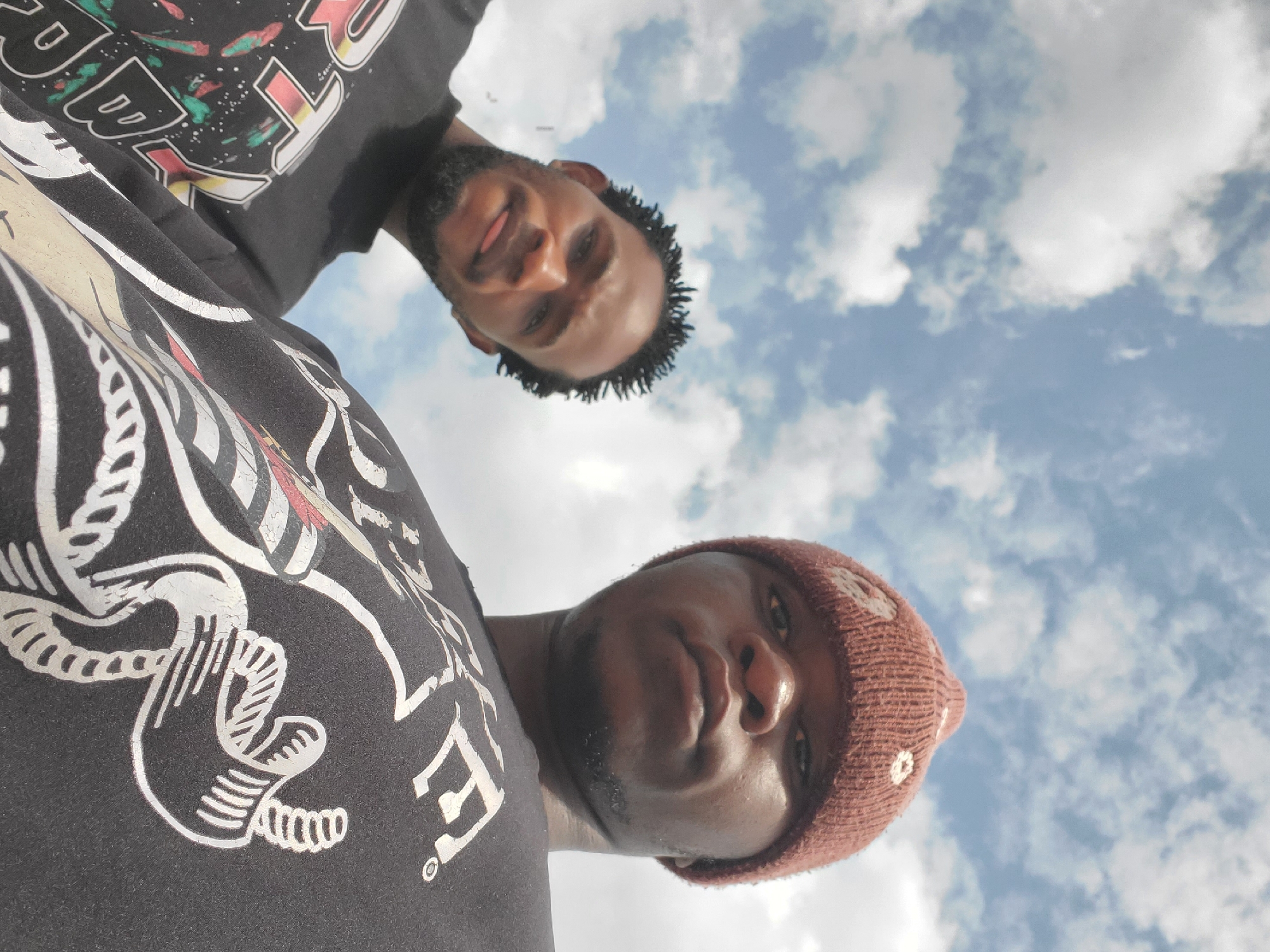 Two young men are captured in a low-angle, close-up selfie against a cloudy blue sky. The man on the right, closer to the camera, wears a brown knit beanie with white floral accents and a black t-shirt featuring a white graphic design that includes what appears to be a skeleton's hand holding a rope. The man on the left, slightly further back and to the left, has dark, braided hair and a goatee. He is wearing a black t-shirt with a colorful graphic design. The image was taken outdoors, possibly in a park or open area, given the sky and lack of urban structures. There is no indication of the city of Winters, United States of America in the image or its context.