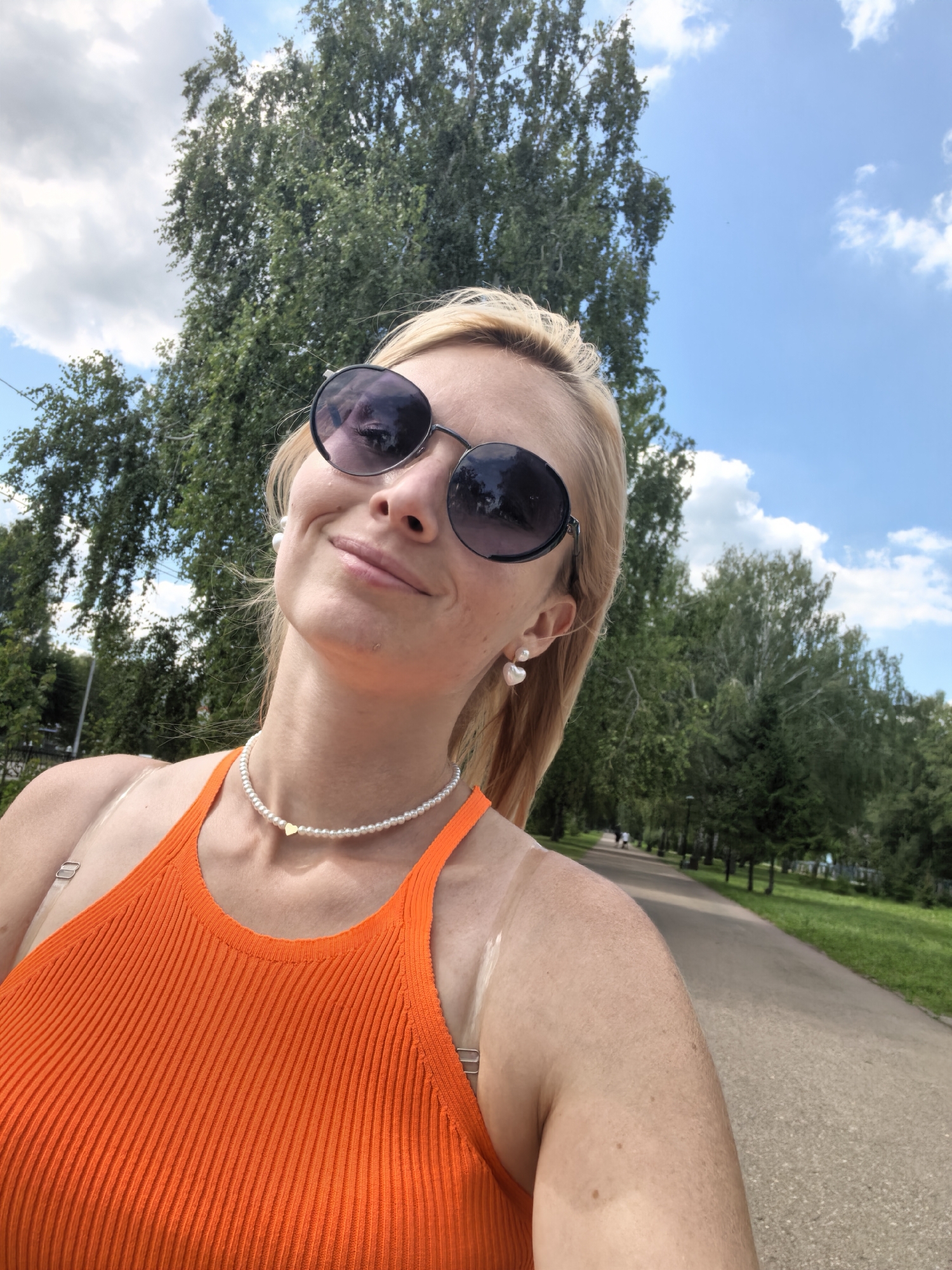 A blonde woman wearing sunglasses and an orange top is taking a selfie in a park. She has on a pearl necklace with a gold heart pendant and heart-shaped earrings. In the background, there are tall trees and a paved path. The sky is blue with white clouds. The location is a park in Sterlitamak, Russia.