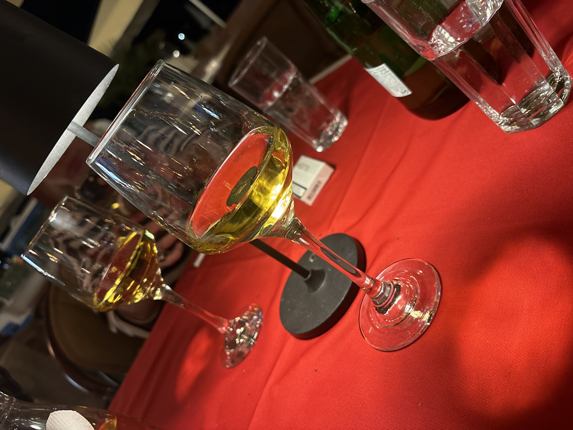 A high-angle shot shows a table covered with a red tablecloth, upon which various drinking glasses are placed. In the foreground are two wine glasses, one filled with a light-colored liquid (likely white wine) and the other partially filled with a similar beverage. A small black lamp base sits between them. Further back, a water glass and a green glass bottle stand out, and a packet of cigarettes or a small box can be seen in between the wine and water glasses. The composition suggests a restaurant or bar setting. The photo doesn't provide information about location in Budva, Montenegro.