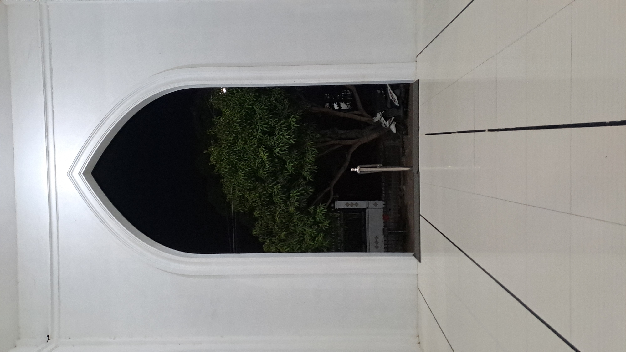 The image shows a view through an arched doorway from an indoor tiled area to an outdoor area at night. The interior features a light-colored tiled floor with dark linear inlays. The doorway is white and has a pointed arch. Outside, a large tree with dense green foliage is visible. The background beyond the tree is dark, suggesting it is nighttime. A light-colored fence or wall with decorative patterns is partially visible behind the tree, and two motorcycles are parked near the base of the tree. A metal pole with a finial is standing in front of the tree. The scene appears to be an outdoor area adjacent to a building, possibly a mosque, given the architectural style of the archway. The location is identified as Lhokawe Teungoh, Indonesia.