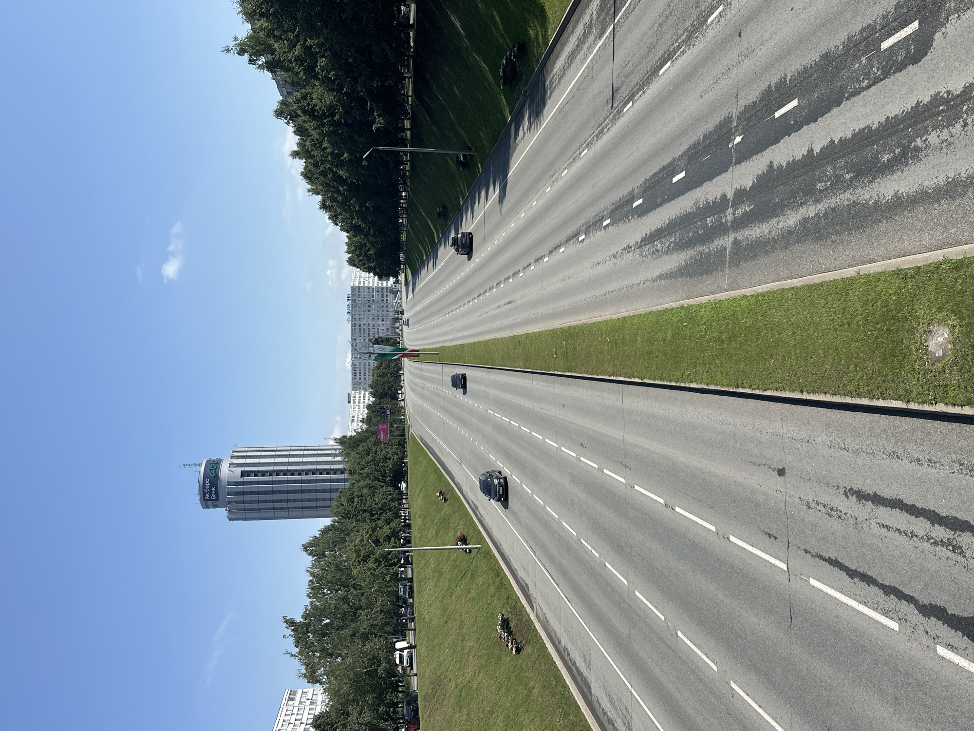 The image shows a wide, multi-lane highway in Naberezhnye Chelny, Russia, under a clear blue sky. The highway is divided by a green median strip. On the left side of the median, there are three lanes of traffic going away from the viewer, and on the right, two lanes of traffic are also moving away. A few cars are visible on the road.
To the left of the highway, there is a grassy embankment with trees and some buildings in the background. A tall, cylindrical building with a sign that reads "АК Барс Банк" (AK BARS BANK) is prominent in the middle ground. On the right side of the highway, another grassy embankment rises, lined with trees, and behind that, more buildings are visible. The road surface shows some signs of wear and dark patches, possibly from oil or water stains. In the distance, more buildings and trees suggest an urban or suburban environment. The overall scene depicts a sunny day in a city with modern infrastructure.