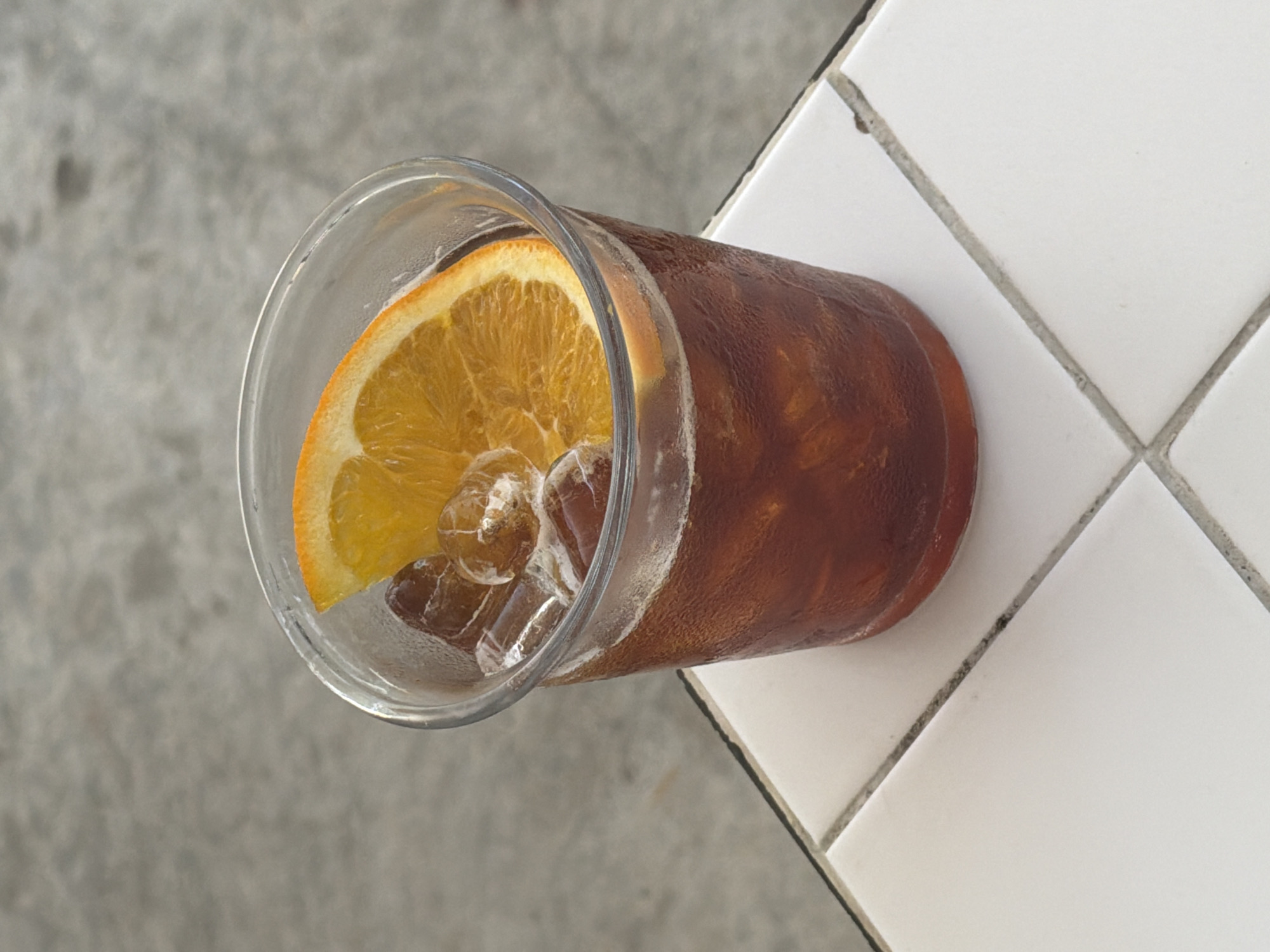 A high-angle shot shows a clear plastic glass filled with a dark brown liquid, likely iced coffee, and garnished with an orange slice and ice cubes. The glass is condensation-covered, indicating its contents are cold. It rests partially on a surface of white square tiles and partially on a rough gray concrete-like surface. The scene's location in Đà Nẵng, Vietnam, is not directly discernible from the image's visible elements.