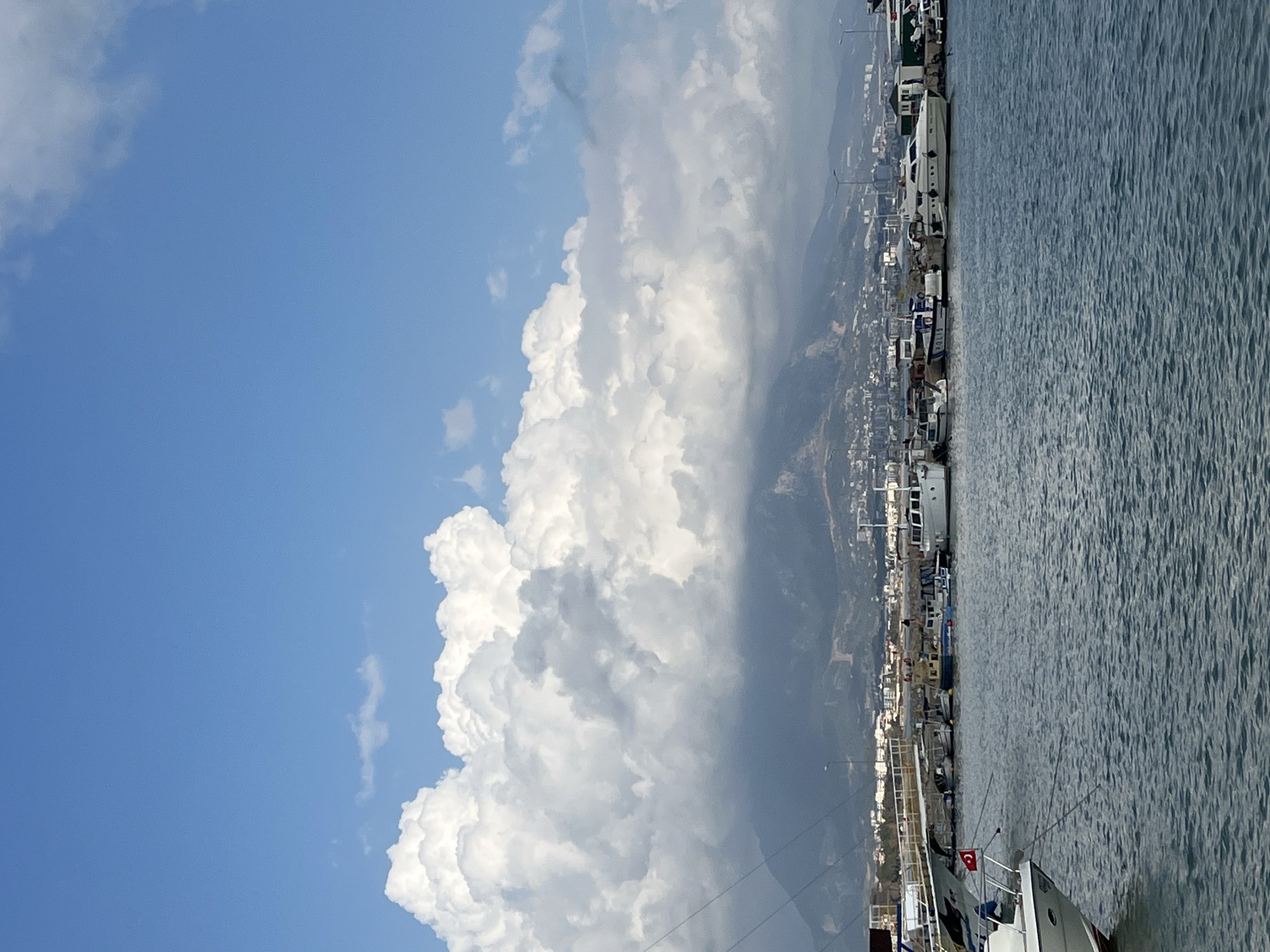 The image shows a view from Alanya, Turkey. The foreground consists of the Mediterranean Sea. Boats, including sailboats, are docked along the harbor. A Turkish flag can be seen on one of the boats. Beyond the harbor, there is a coastal urban area of Alanya, including residential and commercial buildings. A mountain range forms the backdrop to the city. A large cloud formation dominates the left side of the frame and extends toward the city and mountains. A clear blue sky is visible above the clouds.
