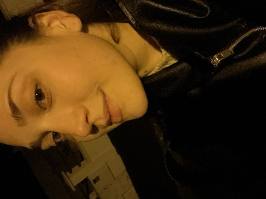 A woman's face is close-up, angled to the left, with an open-mouthed smile. She has fair skin, dark eyebrows, and brown eyes. To her right is a black leather jacket with a visible zipper pull. The background is dark, with a hint of a light-colored wall and structure behind her left side. The image has a warm, yellow cast to it.