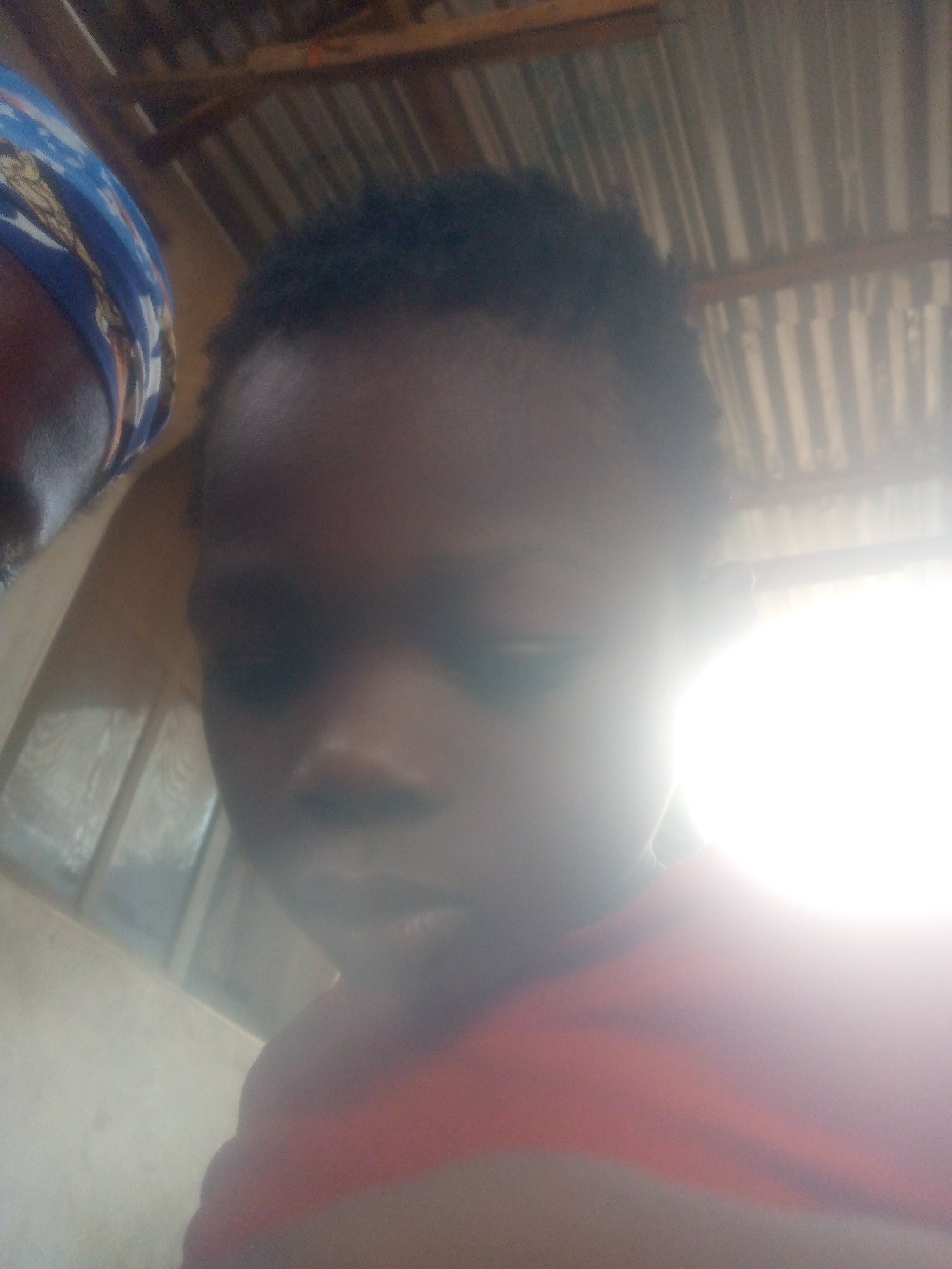 A child's face is partially obscured by a bright light source on the right side of the frame. The child appears to have short, dark hair. Another person wearing a blue headscarf is partially visible on the left side of the frame. The background shows corrugated metal roofing and a wall with a window. The location is in Ikorodu, Nigeria.