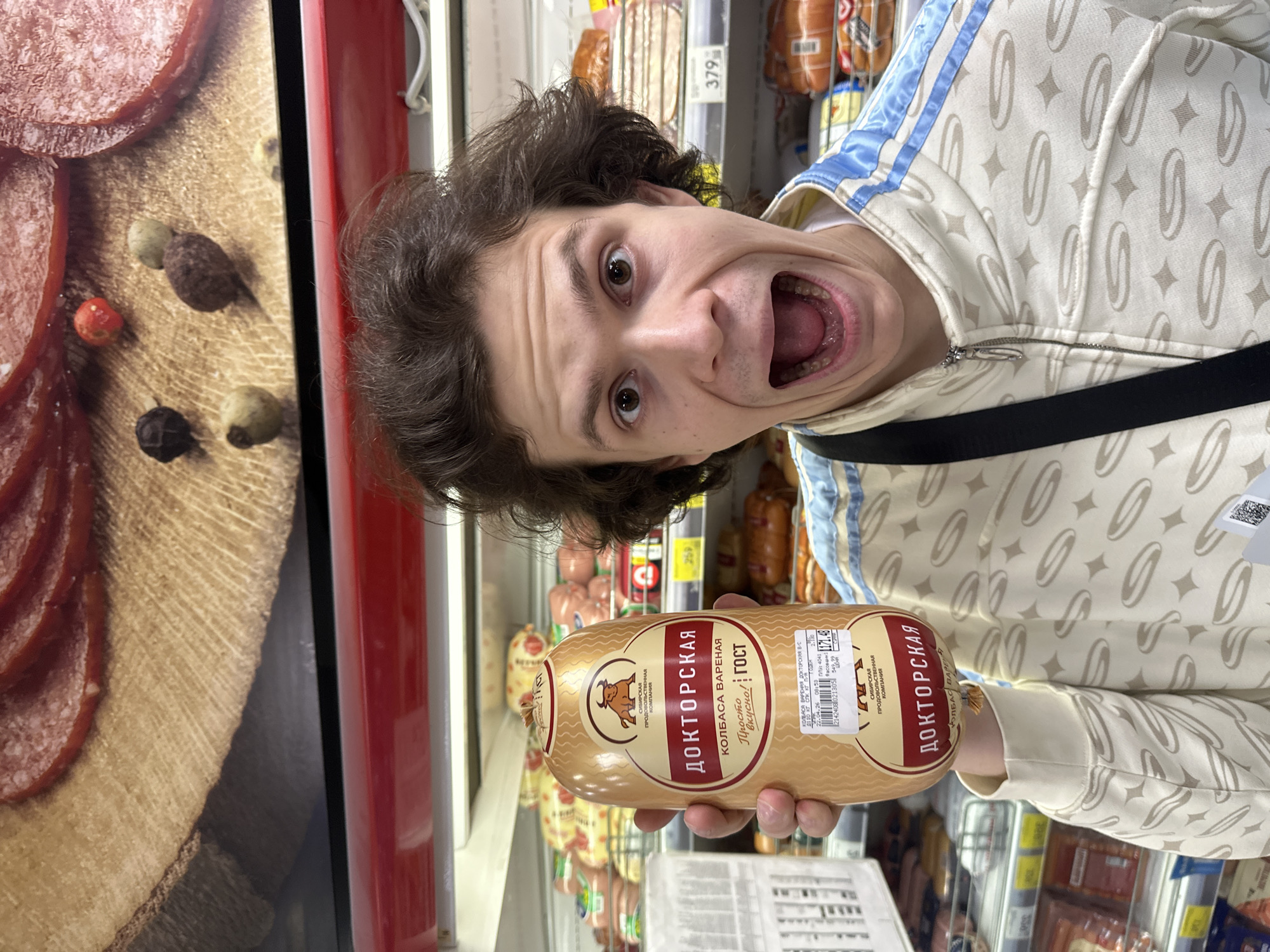 Here's a detailed description of the image:
**General Overview:**
The image shows a person posing comically in what appears to be a grocery store aisle. They are holding up a package of sausage. The setting is likely a supermarket or similar retail food establishment.
**People:**
* There is one person clearly visible. This person is a young adult with dark, curly hair and is making an exaggerated facial expression with their mouth wide open. They are wearing a patterned light-colored jacket with blue stripes and a black strap across their body.
**Subjects and Objects:**
* **Sausage:** The main object is a package of "Doktorskaya" (Докторская) sausage, a type of cooked sausage popular in Russia and other countries of the former Soviet Union. The label indicates that it is a "KOLBASA VARENAYA" (boiled sausage). The package indicates production by "Sibirkaya Prodovolstvennaya Kompaniya". There is a price label showing a weight of 549.99 and a price of 171.48.
* **Store Shelves:** The background shows shelves stocked with various types of sausages and other meat products. There are yellow price tags visible on the shelves. One visible price is 379.
* **Advertisement:** To the left, there is an advertisement displaying a picture of salami and peppers.
**Actions and Interactions:**
* The primary action is the person holding the sausage package and posing for the camera with a wide-open mouth. The intention appears to be humorous.
* The person is presumably in the act of shopping or has just selected the sausage.
**Environment:**
* The environment is a typical grocery store aisle. The shelves are well-stocked, suggesting a modern retail setting.
**Other details:**
* The sausage package label contains Cyrillic text, indicating that the image was taken in a region where the Russian language is common.
It's difficult to pinpoint the exact location as the picture doesn't provide enough context. However, based on the sausage type, Cyrillic text, and the company name, it is safe to assume the location is in Russia, more specifically in Krasnoyarsk.