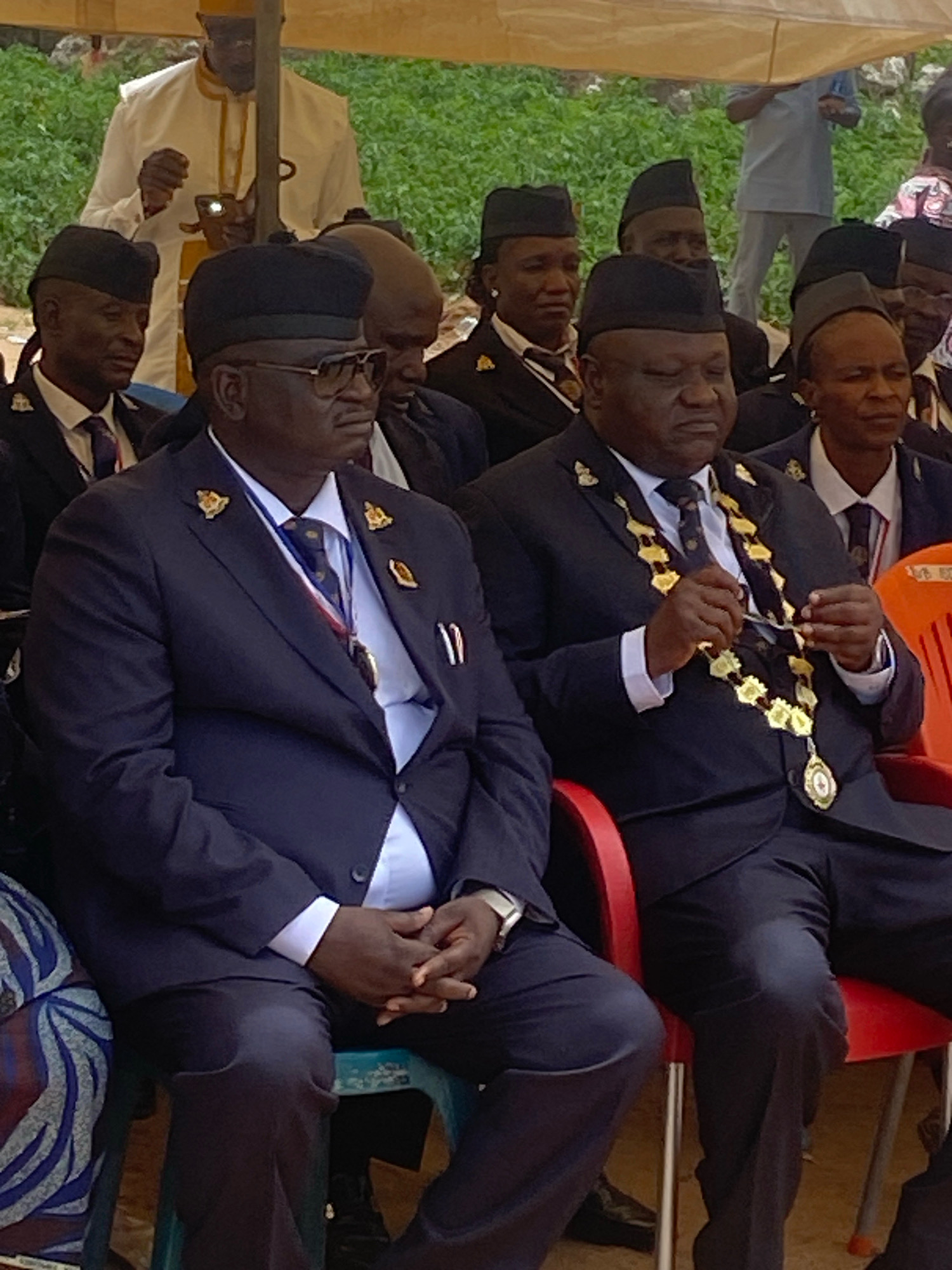 In Binchi, Nigeria, a group of men in formal attire are seated outdoors. The central figure is a Black man wearing a dark suit, a white shirt, a patterned tie, and a ceremonial gold chain with a pendant. He is seated on a red chair and has a relaxed posture. To his left, another Black man in a dark suit, white shirt, and tie is visible. He wears dark-rimmed glasses and a black cap. To the right of the central figure, another man in a similar dark suit and white shirt is seated, his face mostly obscured.
In the background, more individuals are present, some wearing black caps. One man in the upper background is wearing a yellow and white robe and a yellow cap, holding a small metallic object. The setting appears to be under a light-colored canopy, with greenery visible behind the seated individuals. The ground is partially visible, showing a mix of earth and possibly some worn fabric or plastic sheeting. The overall atmosphere suggests a formal gathering or ceremony.
