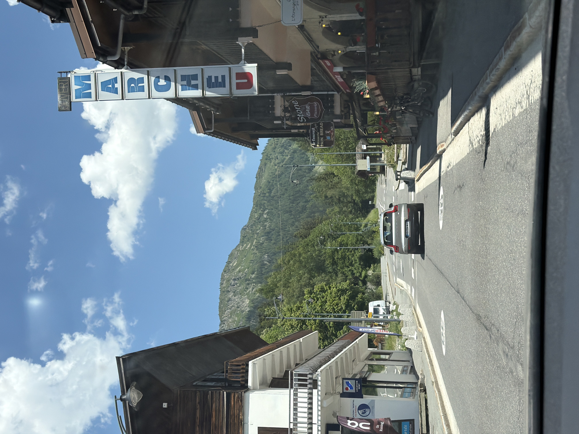 The image captures a sunlit street in Vallorcine, France, on a clear day with fluffy white clouds against a bright blue sky. The street is paved, with white lane markings and speed limit signs indicating "30". On the left side of the street, a multi-story building features a wooden facade on its upper levels and white painted lower levels with balconies. Shops are visible on the ground floor, with signage for "ON Running" and "LOCATION RENT". On the right side, a vertical sign with blue lettering spells out "MARCHE" ("Market") with "OUVERT" ("Open") at the top. Further down the street, more buildings with wooden exteriors house businesses including a "Stone BAR PIZZERIA" and a "L'Étranger" establishment. A car is driving away from the camera down the street, and on the right sidewalk, bicycles are parked outside a wooden establishment. Lush green trees and a forested mountain slope form the backdrop to the scene.