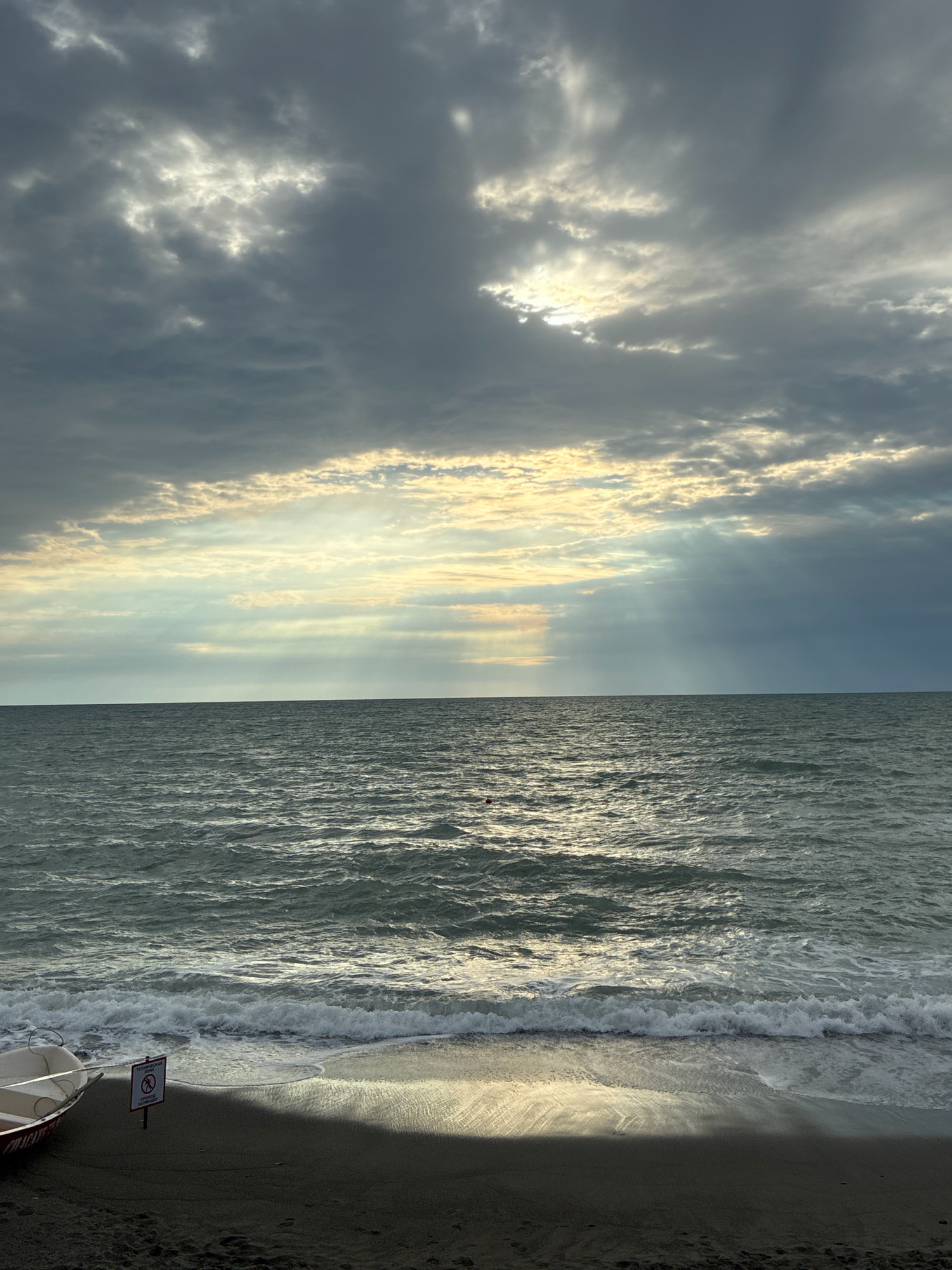 The image captures a serene beach scene on the coast of Sochi, Russia, under a dramatic, cloud-filled sky. The sun's rays pierce through the clouds, casting ethereal shafts of light onto the rippling, dark water of the Black Sea. Gentle waves lap the sandy shore, leaving a glistening wet sand line. In the foreground, a small, red and white boat rests on the dark sand. To its right, a red and white sign on a pole indicates a restricted area. The overall atmosphere is one of calm, with the play of light and shadow creating a moody and picturesque setting.