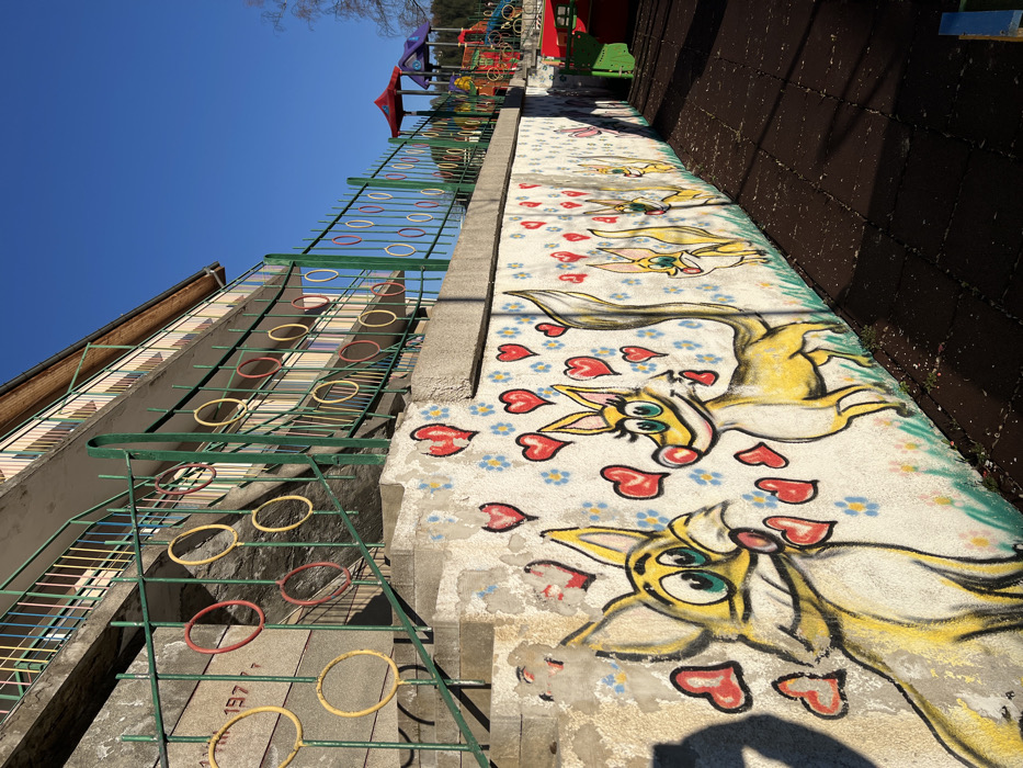 The image depicts an outdoor scene in the city of Banite, Bulgaria, featuring a building, a retaining wall with a mural, and a children's playground under a clear blue sky.
On the upper left, a multi-story, light-colored concrete building, likely a public institution, displays a partially visible "197" embossed on its facade. The building features balconies with light wood soffits and railings. A concrete staircase ascends along the building's side, fitted with a dark green metal railing. This railing transitions into a horizontal dark green metal fence adorned with alternating yellow and red circular decorative rings.
Below the fence, a concrete retaining wall spans horizontally, separating the elevated area from the lower ground. This wall is covered with a colorful mural. The mural depicts multiple cartoon-style, yellow, anthropomorphic creatures resembling foxes or dogs, characterized by large ears, green eyes, and smiling expressions. These figures are interspersed with numerous red heart shapes of various sizes and scattered patterns of small blue and white five-petal flowers against a white background, with a green border representing ground.
To the right, at a lower elevation, a children's playground is visible. The playground's surface consists of dark brown interlocking rubber tiles. Visible equipment includes multi-colored structures featuring slides and platforms with red and blue roofs.
The scene is well-lit by natural sunlight, casting shadows on the mural and playground surface. The sky overhead is clear and bright blue. Sparse, bare tree branches are partially visible on the far right.