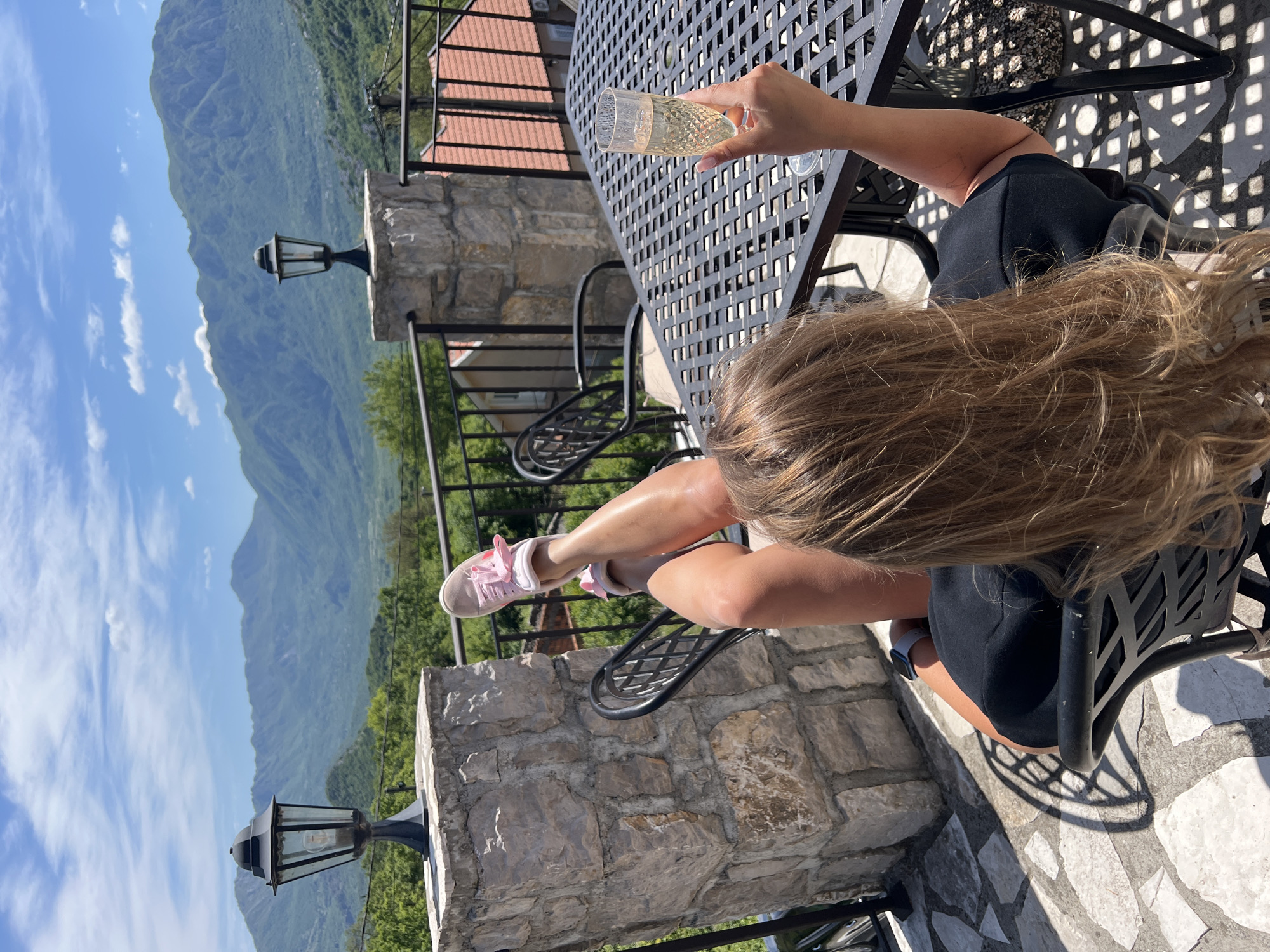 A woman sits at a black metal table on a stone terrace in Sotonići, Montenegro. She holds a glass of champagne and wears a black dress, a grey smart watch, and pink sneakers. The mountains of Montenegro are visible in the background with a blue sky with thin white clouds. A black metal railing and stone pillars with lamp posts are on the terrace.