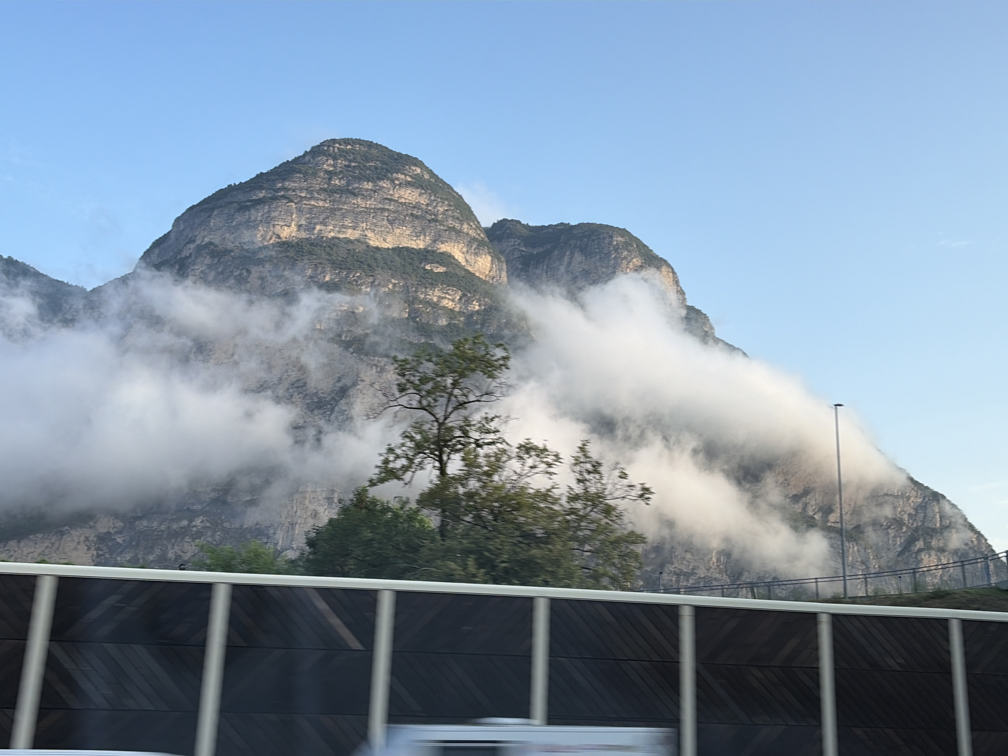 The image captures a majestic mountain range partially obscured by clouds, set against a clear blue sky. The mountains exhibit layered rock formations and are covered with sparse vegetation. In the foreground, a dark, slatted sound barrier runs horizontally across the frame, with vertical white posts. A street lamp is visible on the right side, positioned near the top of the mountain. The scene appears to be on the outskirts of Salorno sulla Strada del Vino - Salurn an der Weinstraße, Italy, given the typical infrastructure seen along major roadways in mountainous regions.