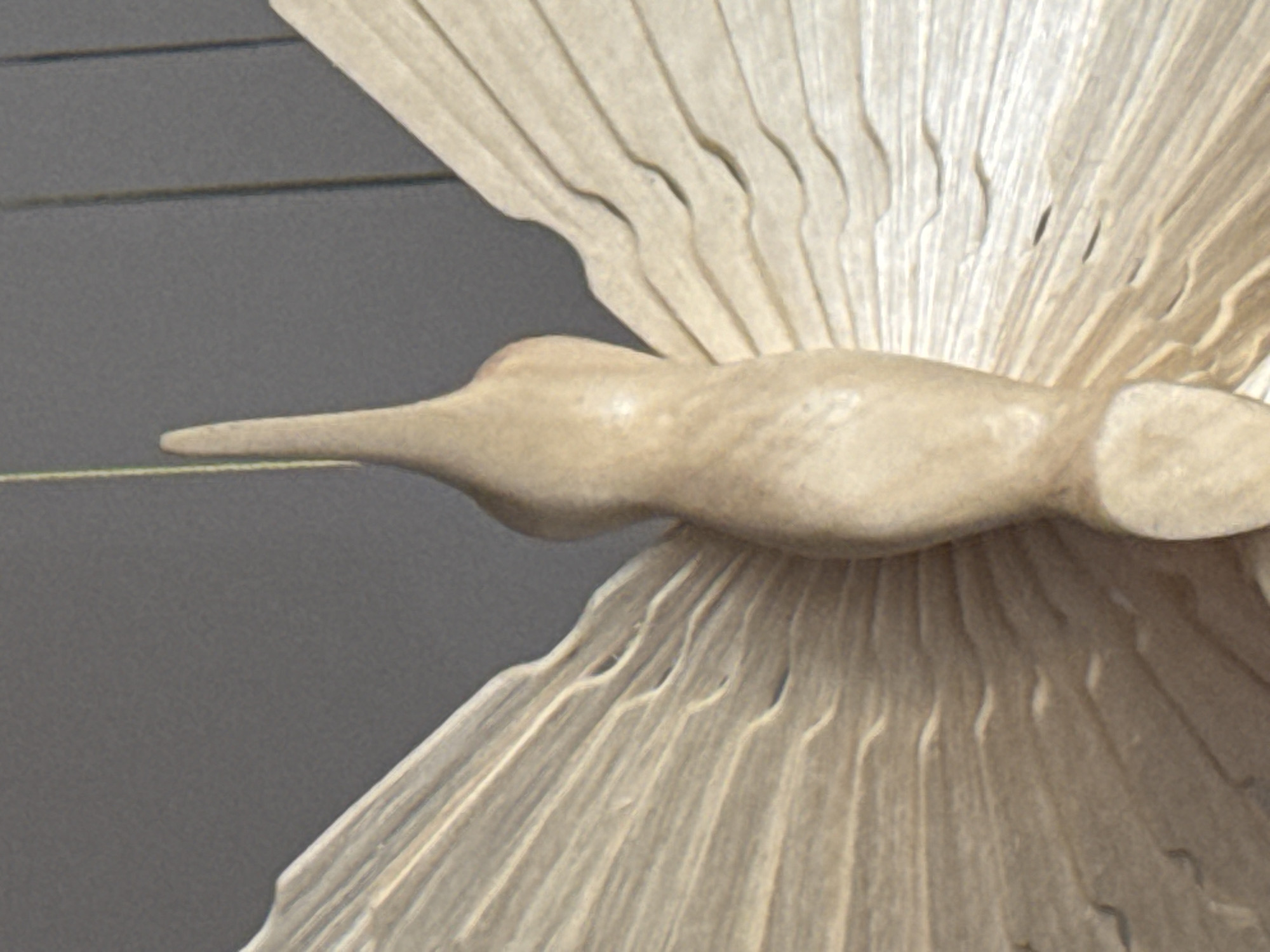 A close-up, eye-level, medium shot captures a light brown, wooden butterfly sculpture with its wings spread wide. The sculpture is viewed from the back, with its elongated body pointing upwards and a slender antenna extending from its head. The wings, intricately carved with delicate lines resembling veins, fan outwards on either side of the body. The background is a plain, dark gray wall, possibly a room interior, which is out of focus, drawing all attention to the sculpted butterfly. There is no indication of the city of Kholmsk, Russia, or any specific location within it.