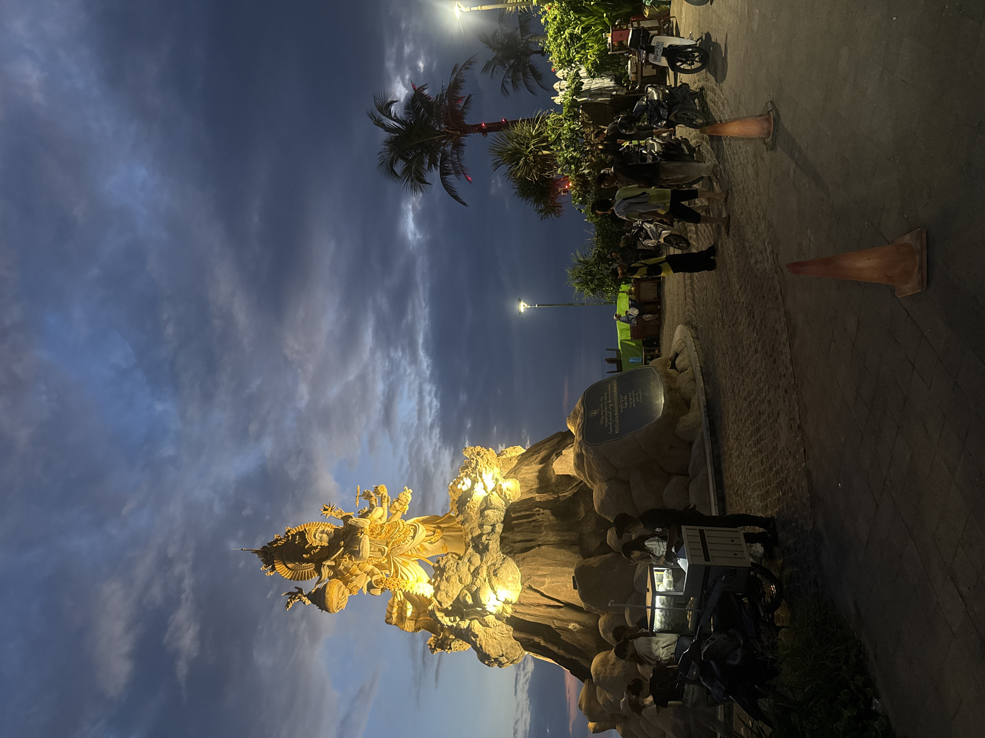 The image shows a view of a statue in North Kuta, Indonesia, at night. The statue is brightly illuminated and appears golden against the backdrop of a cloudy evening sky. Nearby are palm trees, some decorated with red lights.
People are gathered around a vendor or food cart in the statue's vicinity. Several motorbikes are parked nearby. Orange traffic cones are also visible on the walkway.