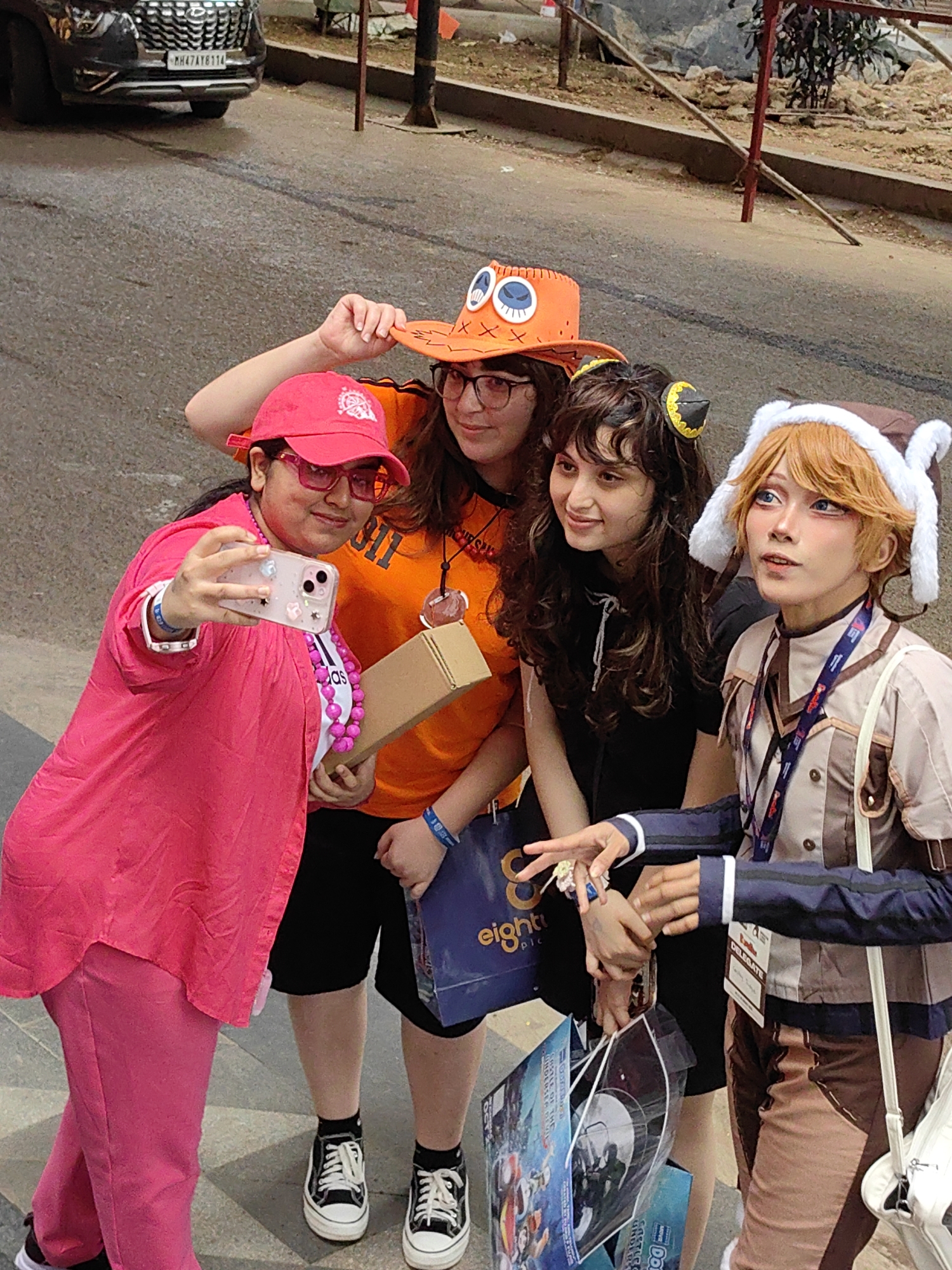 Four individuals, likely attending an event, are posing for a photograph on a paved city street. The person on the far left, wearing a bright pink outfit and hat, is holding up a smartphone to take a selfie. The individual to their right wears an orange shirt and a distinctive orange hat with eye details. The third person is dressed in black and has dark, curly hair. The person on the far right is wearing a costume with blonde hair, blue-tinted contact lenses, and a brown hat with white fluffy ear-like decorations. They are all grouped closely together. Behind them, a dark-colored car with the license plate "MH47AT8114" is visible parked on the street, indicating the scene is in Mumbai, India, as the "MH" prefix denotes Maharashtra. The ground is wet in places, suggesting recent rain or a damp environment. The background shows some construction or urban development with scaffolding and debris.