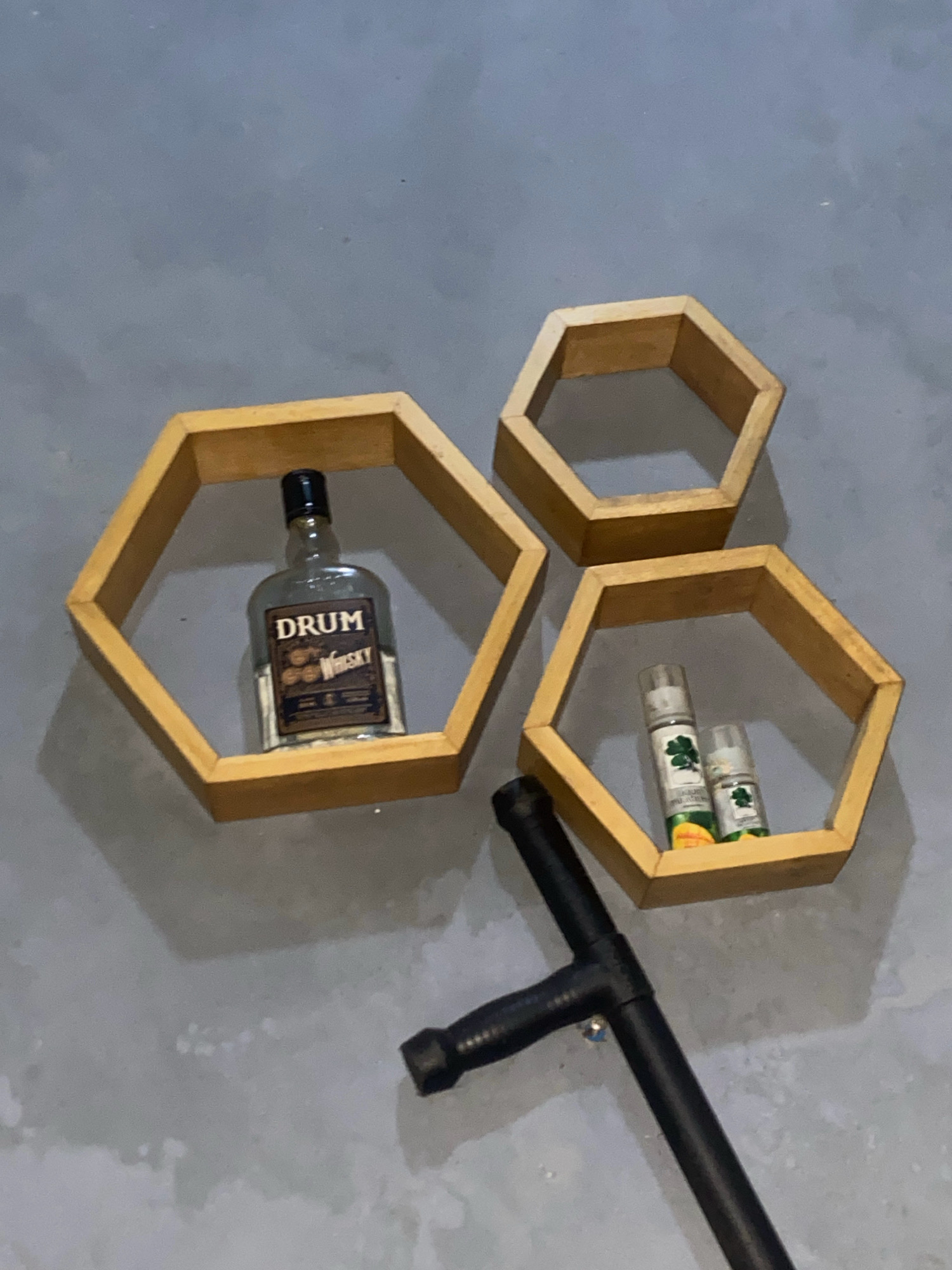 Three hexagonal wooden shelves are mounted on a gray, textured wall. A bottle of "DRUM" whisky is displayed on the largest, left-hand shelf. Two smaller bottles, possibly personal care items with green leaf imagery, are placed on the smallest, lower-right shelf. Below the shelves and to the right, a black baton is resting on the floor. The scene appears to be an indoor setting, potentially a residential or commercial space, in Batam, Indonesia.