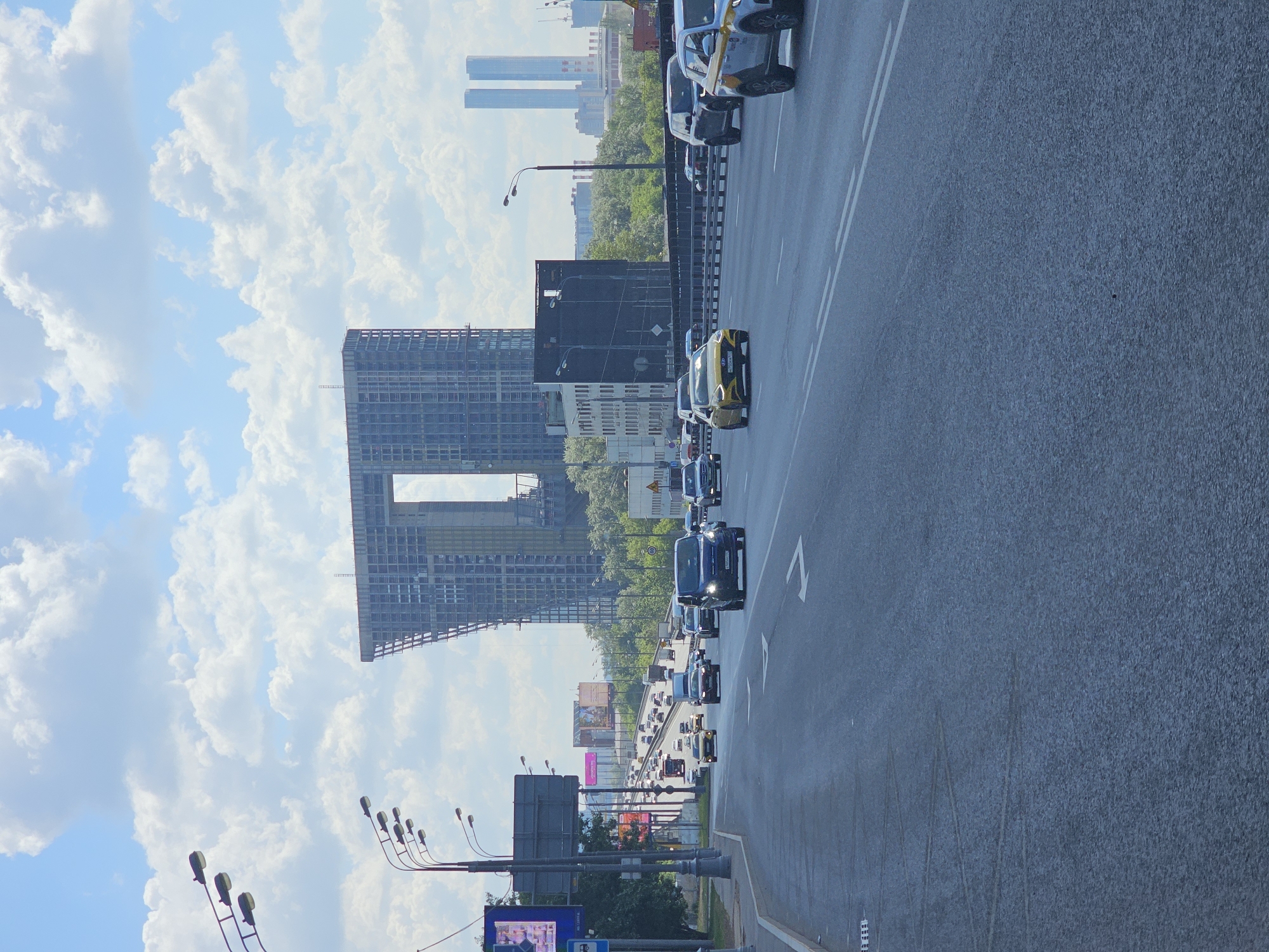 A daytime view captures a bustling multi-lane highway or major road in Moscow, Russia, under a partly cloudy sky. The road, which dominates the right side of the frame, shows a continuous flow of cars, including several with yellow markings, likely taxis. White road markings, including solid lines, dashed lines, and directional arrows, are visible on the asphalt surface.
To the left of the road, the urban landscape unfolds. A large, distinctive modern building, partially constructed and featuring a prominent, large rectangular opening in its upper section, stands out. Adjacent to it, a blockier, lighter-colored building can be seen. Further in the background, beyond a stretch of green trees, several distant, taller skyscrapers are visible against the horizon.
Streetlights on dark poles line the side of the road, and various billboards, some displaying colorful advertisements, are scattered throughout the scene, though their text is largely unreadable. A blue sign below a streetlight on the left is also too indistinct to decipher. On one of the yellow taxis, part of a Russian license plate, "M 109," is discernible. The weather appears bright and clear, indicating a sunny day with scattered white clouds across a blue sky. The scene depicts everyday urban traffic and development.
