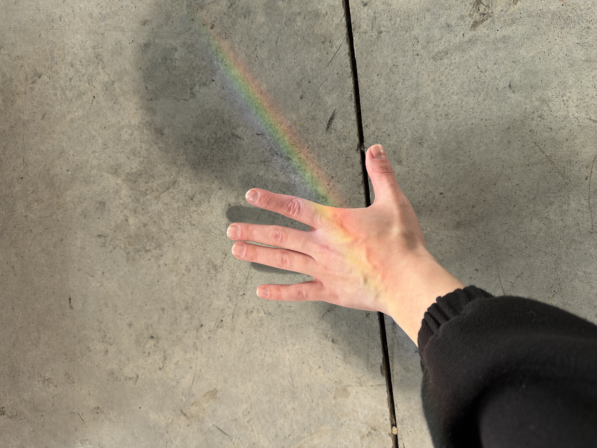 A hand is illuminated by a rainbow-colored light, likely a light refraction effect, on a gray concrete floor. The person is wearing a black long-sleeved top. The floor has a distinct seam or line running vertically down the right side. There is no information available that would indicate this photo was taken in Tel-Aviv, Israel.