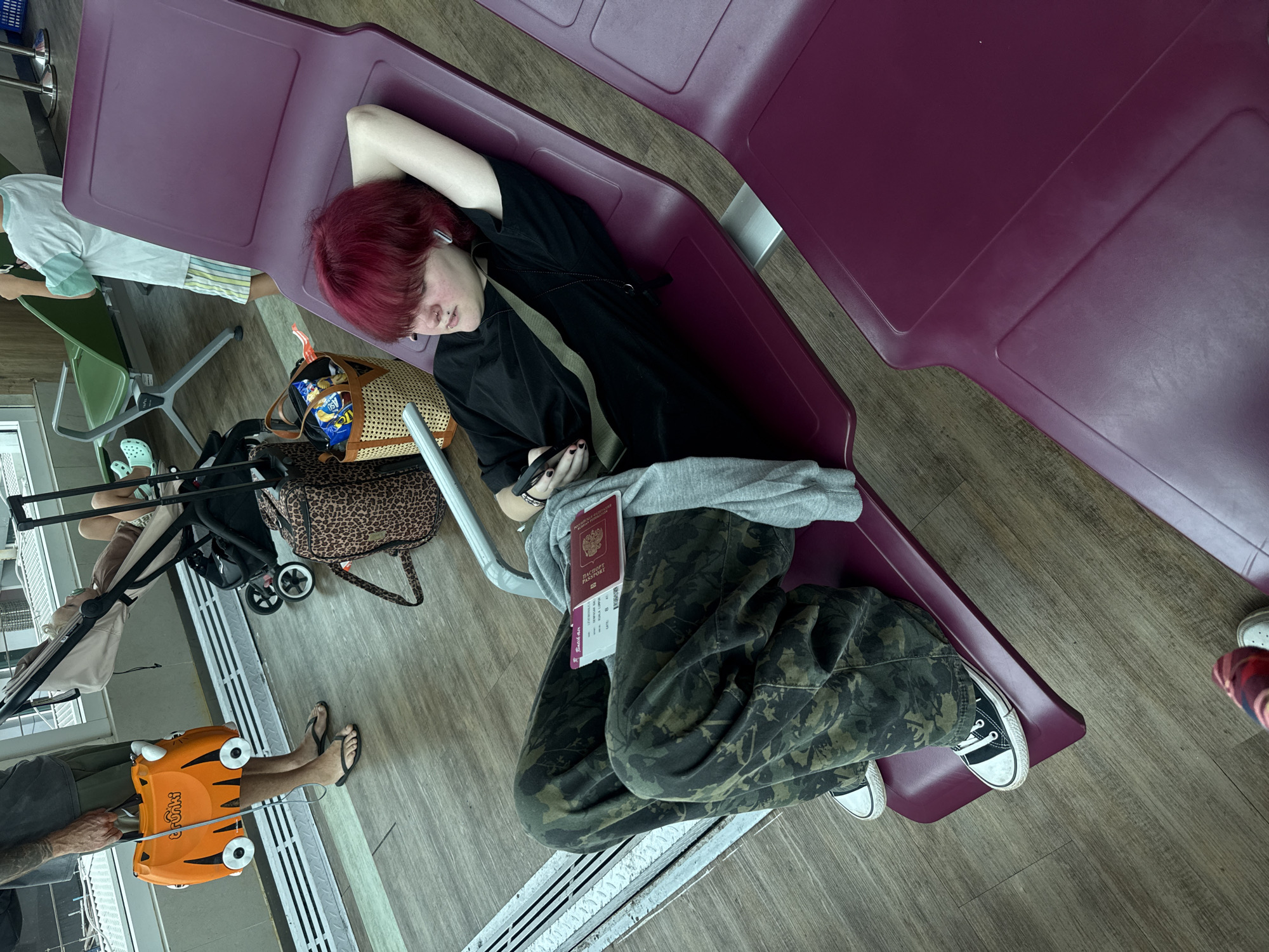 An individual with red hair is lying on a purple airport lounge seat, with their head resting on their hand. They are wearing a black shirt, grey sweater, and camouflage pants, and have a Russian passport on their lap. An orange Trunki suitcase is on the floor near a person wearing sandals. A bag with snacks is on the floor near a baby stroller. The location is potentially an airport in Kuta, Indonesia.